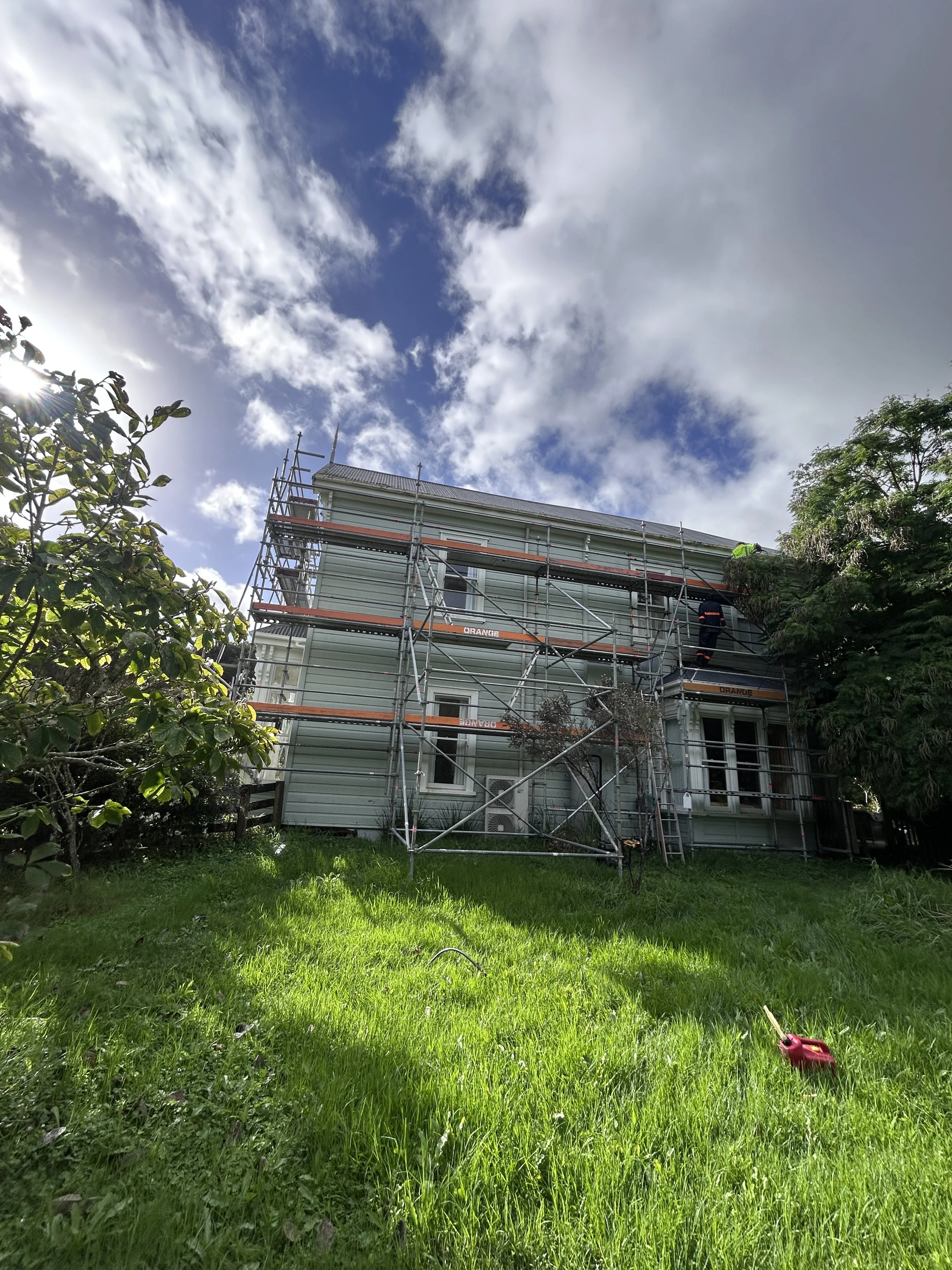A two-story house under renovation surrounded by green grass and trees, with scaffolding set up around it, and workers on site. The sky is partly cloudy with patches of blue.