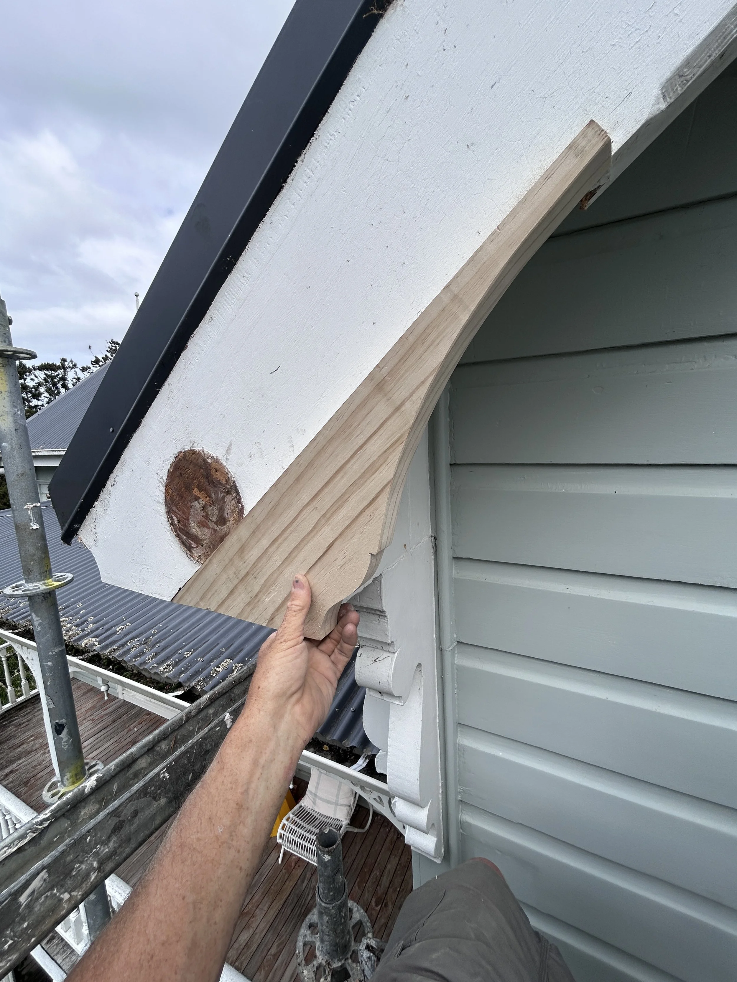 Person holding a curved piece of unfinished wood siding next to a white house wall under construction or repair, with scaffolding and part of a deck visible in the background.