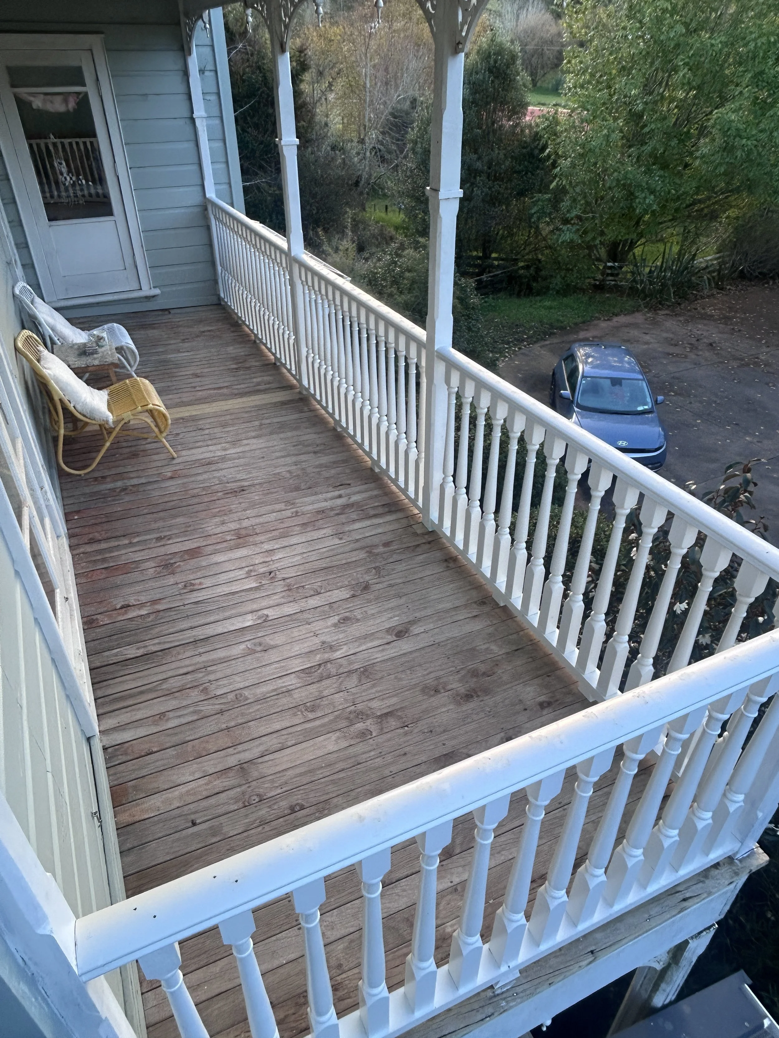 A wooden balcony with white railing and a white lattice top, with a view of trees, a parked gray car, and a dirt area below.