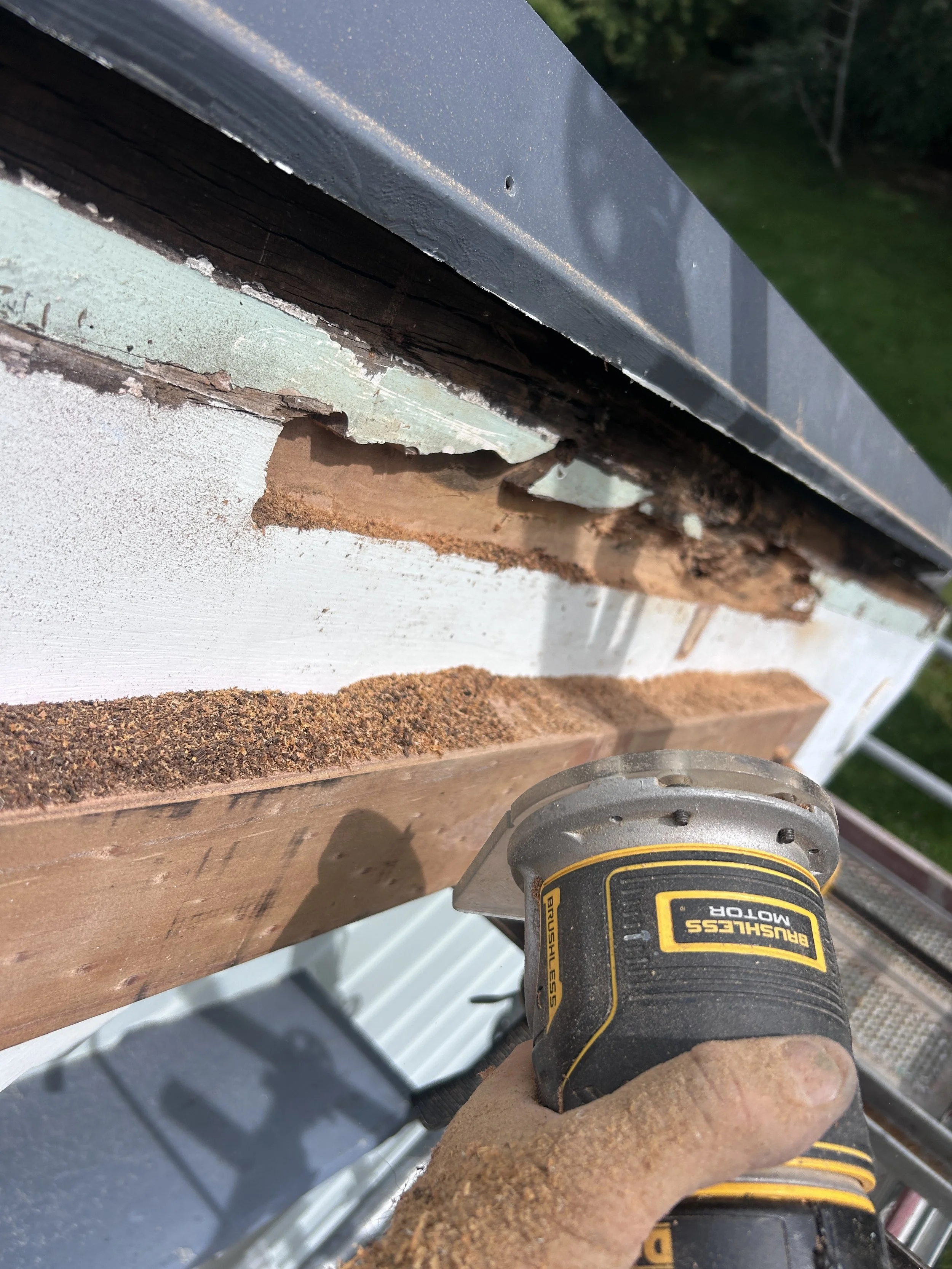 Close-up of a person's hand using a rotary tool on a wooden surface, with visible sawdust and a metal structure in the background.