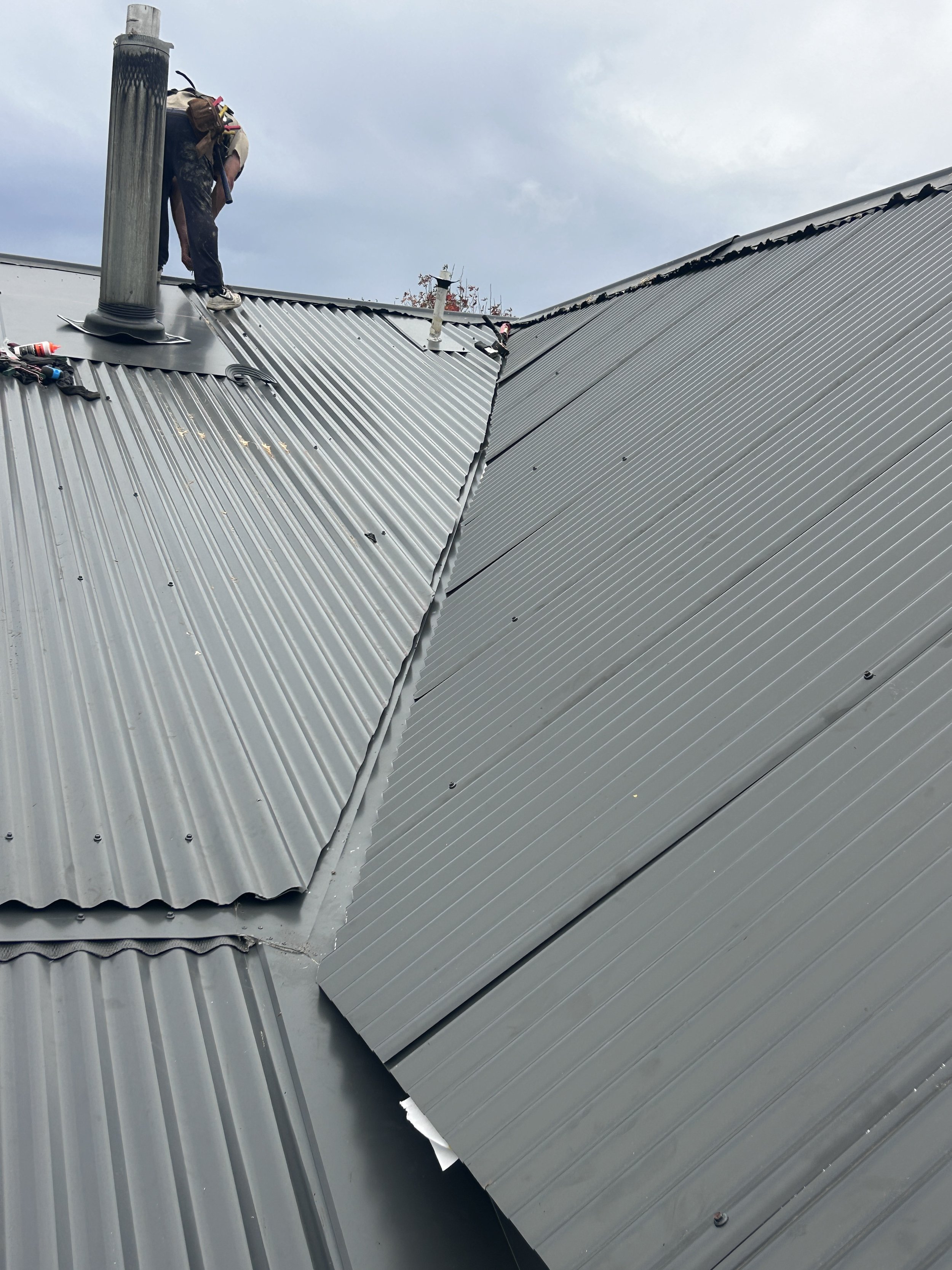 A worker on a metal roof installing or repairing the corrugated metal panels, equipped with tools and safety gear, under a cloudy sky.
