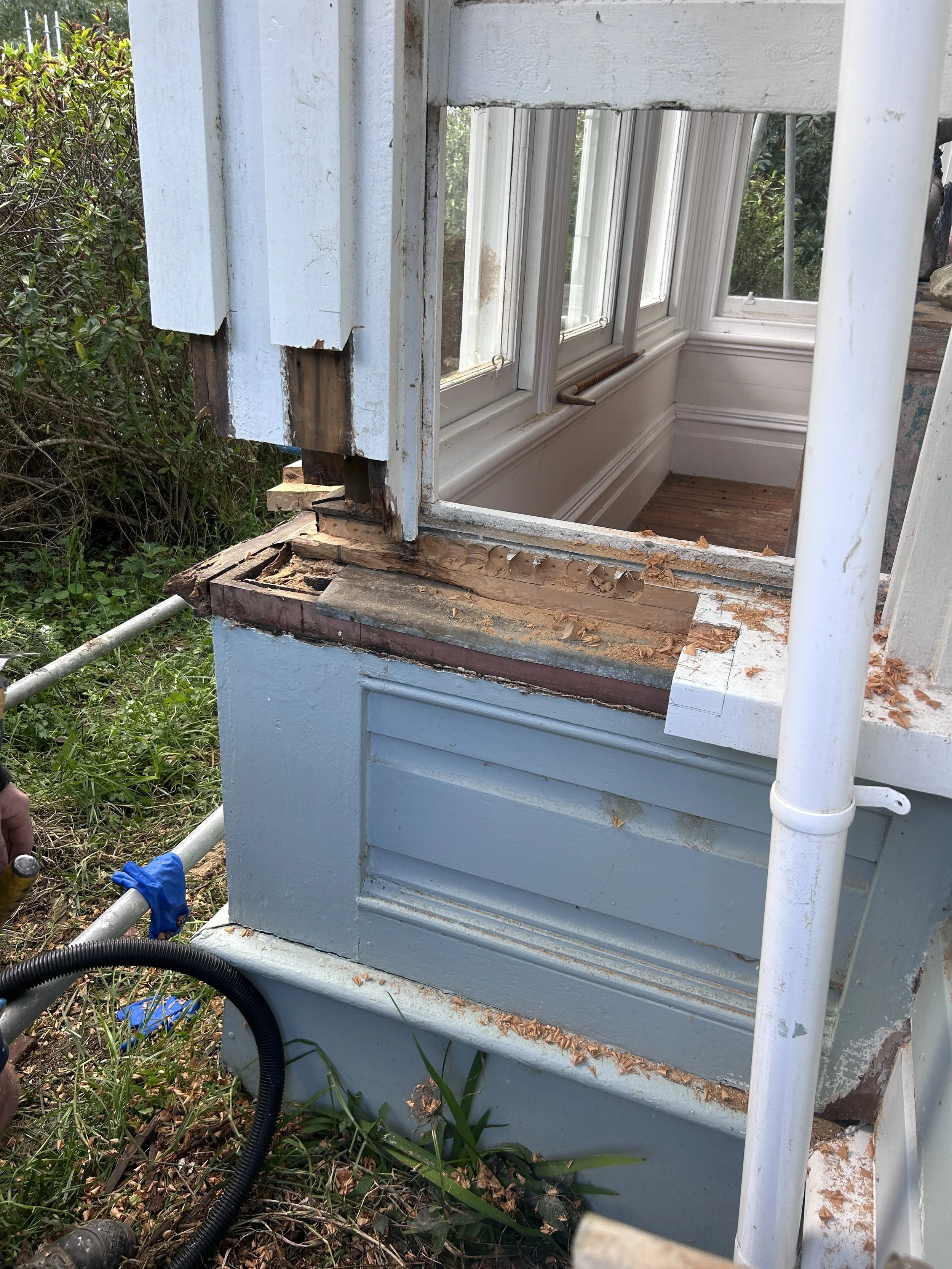 Partial view of a damaged light blue wooden house or structure with exposed wood and peeling paint, surrounded by grass and bushes, with construction tools and pipes nearby.