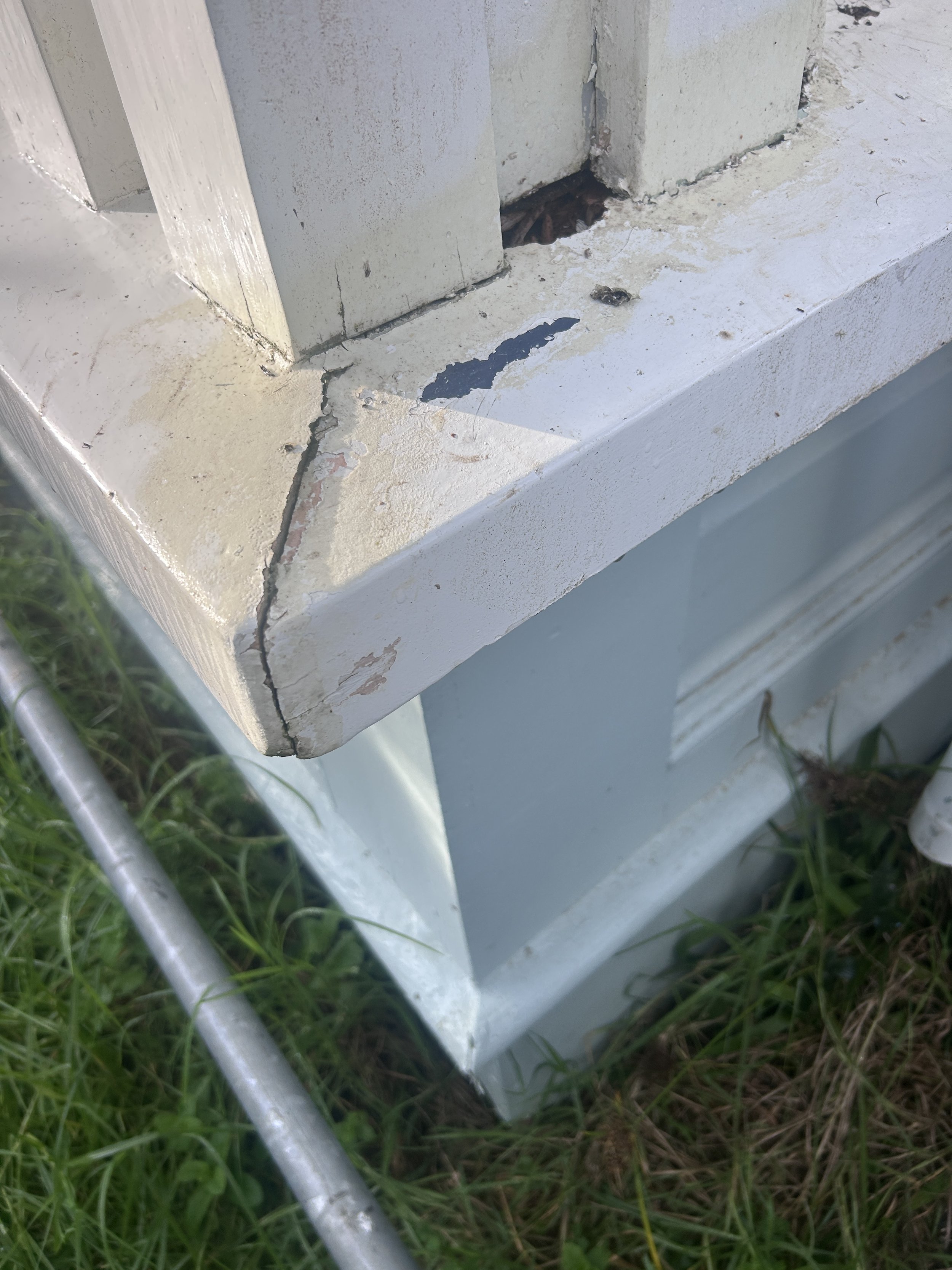 A close-up of a damaged white wooden step with a large crack and chipped paint, situated next to green grass and a silver metal pipe.