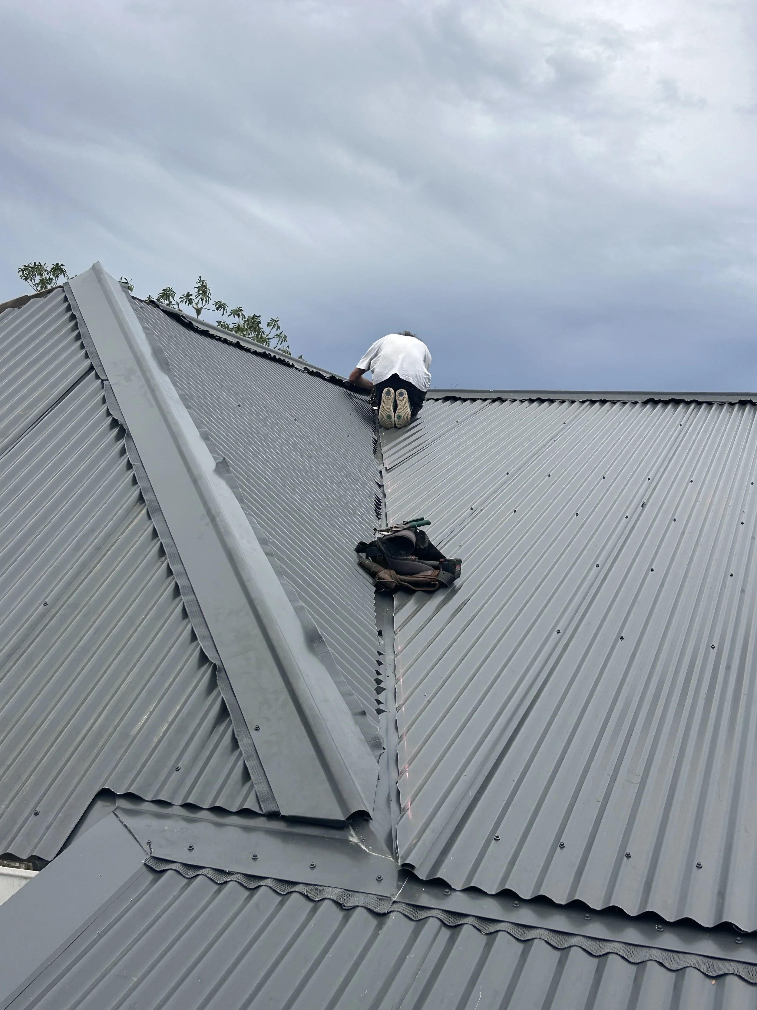 A person working on a metal roof during cloudy weather, kneeling near the peak with tools and a bag nearby.