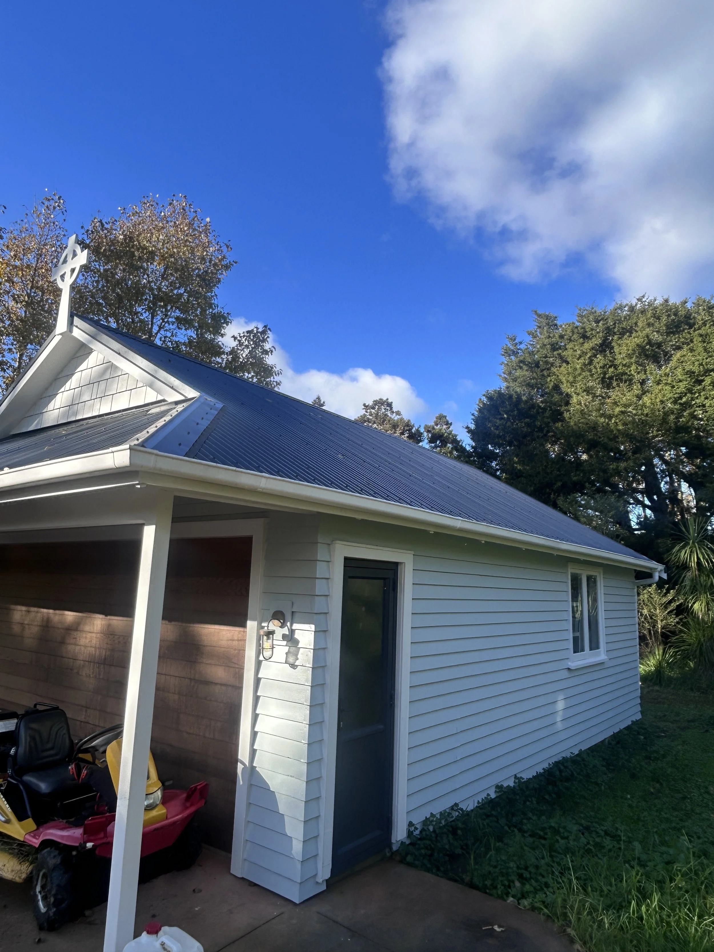 Photo of a white house with a metal roof, a porch area, and a large window. There are trees in the background and a partly cloudy sky. A small storage shed with white siding is attached to the house, with children's toy cars parked underneath.