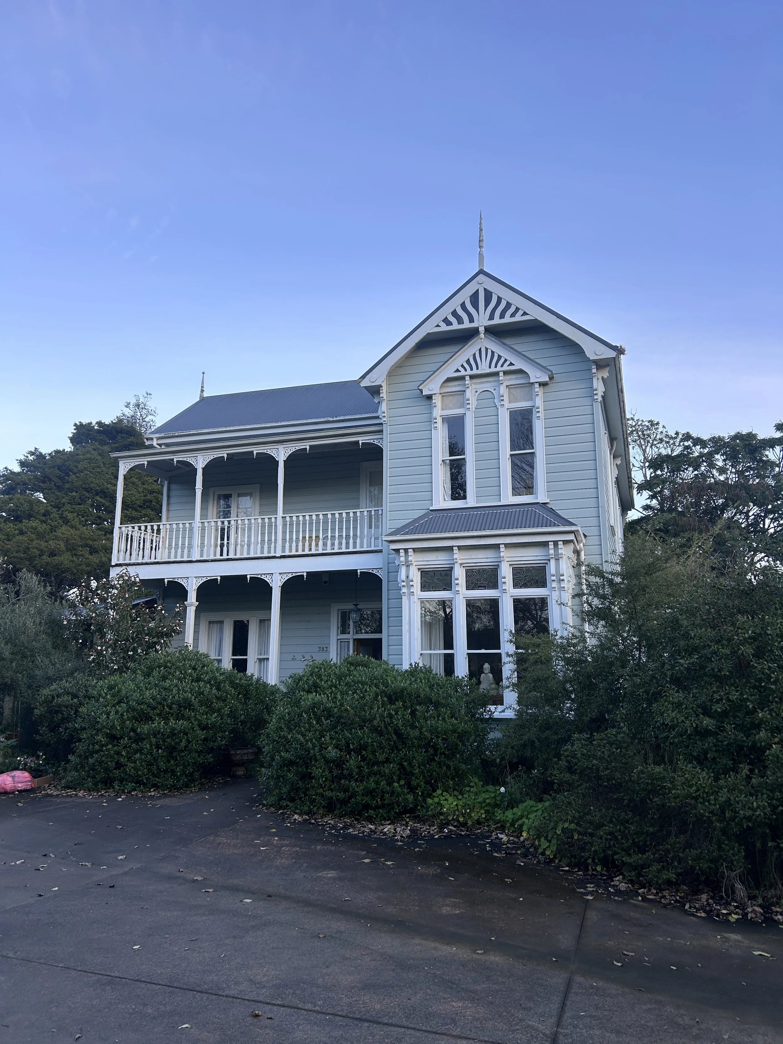 A light blue Victorian style house with decorative trim, large windows, and a wraparound porch, surrounded by greenery, under a clear blue sky.