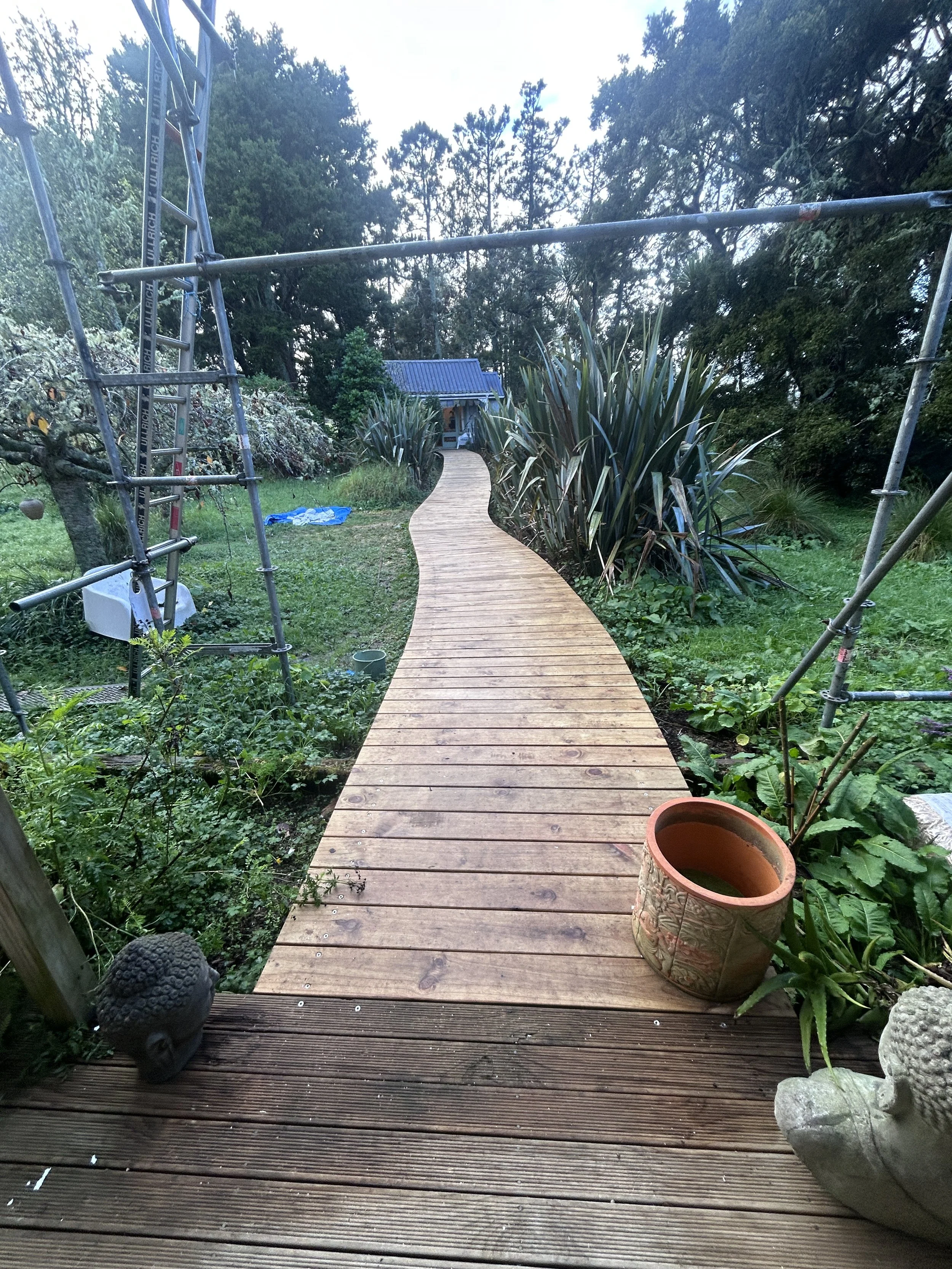 A wooden deck with a curved walkway leading to a blue-roofed structure surrounded by green plants and trees, with construction scaffolding on the sides.