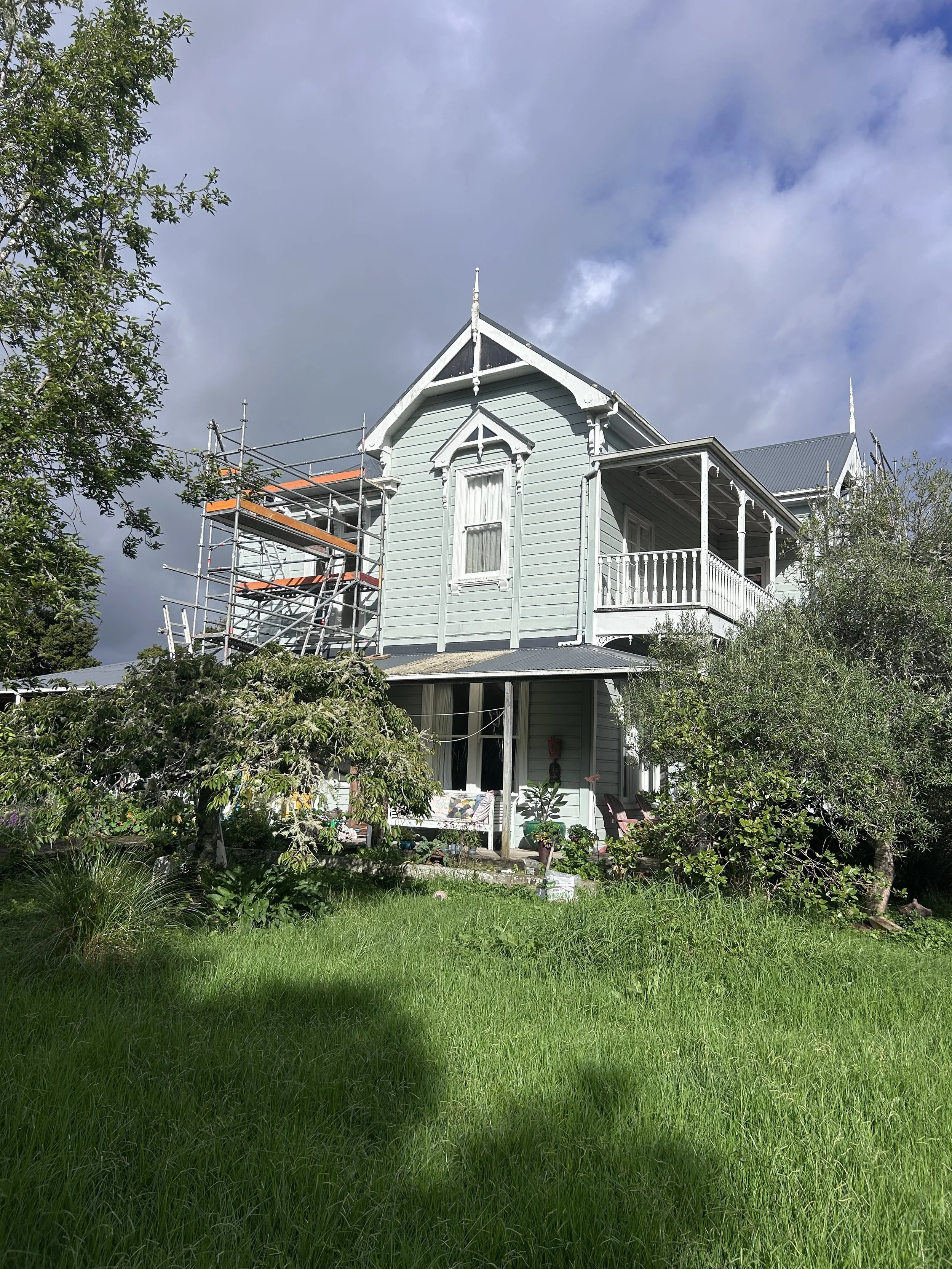 A light blue, two-story house with white trim, a balcony, and scaffolding on the side, surrounded by green trees and grass, under a partly cloudy sky.