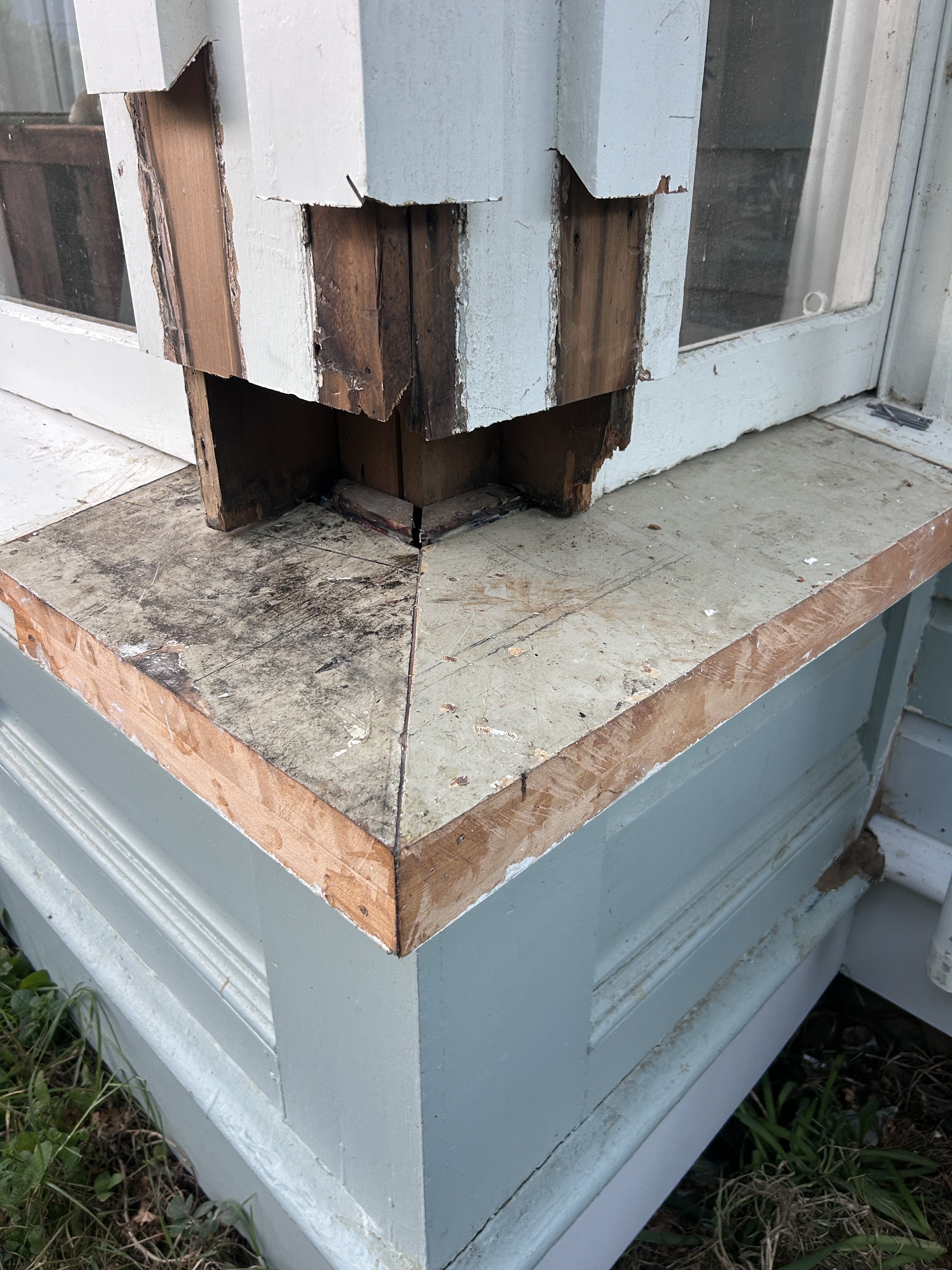 Close-up of a wooden structure with some parts damaged or rotting, placed on a painted surface near a glass door, with grass visible at the bottom.