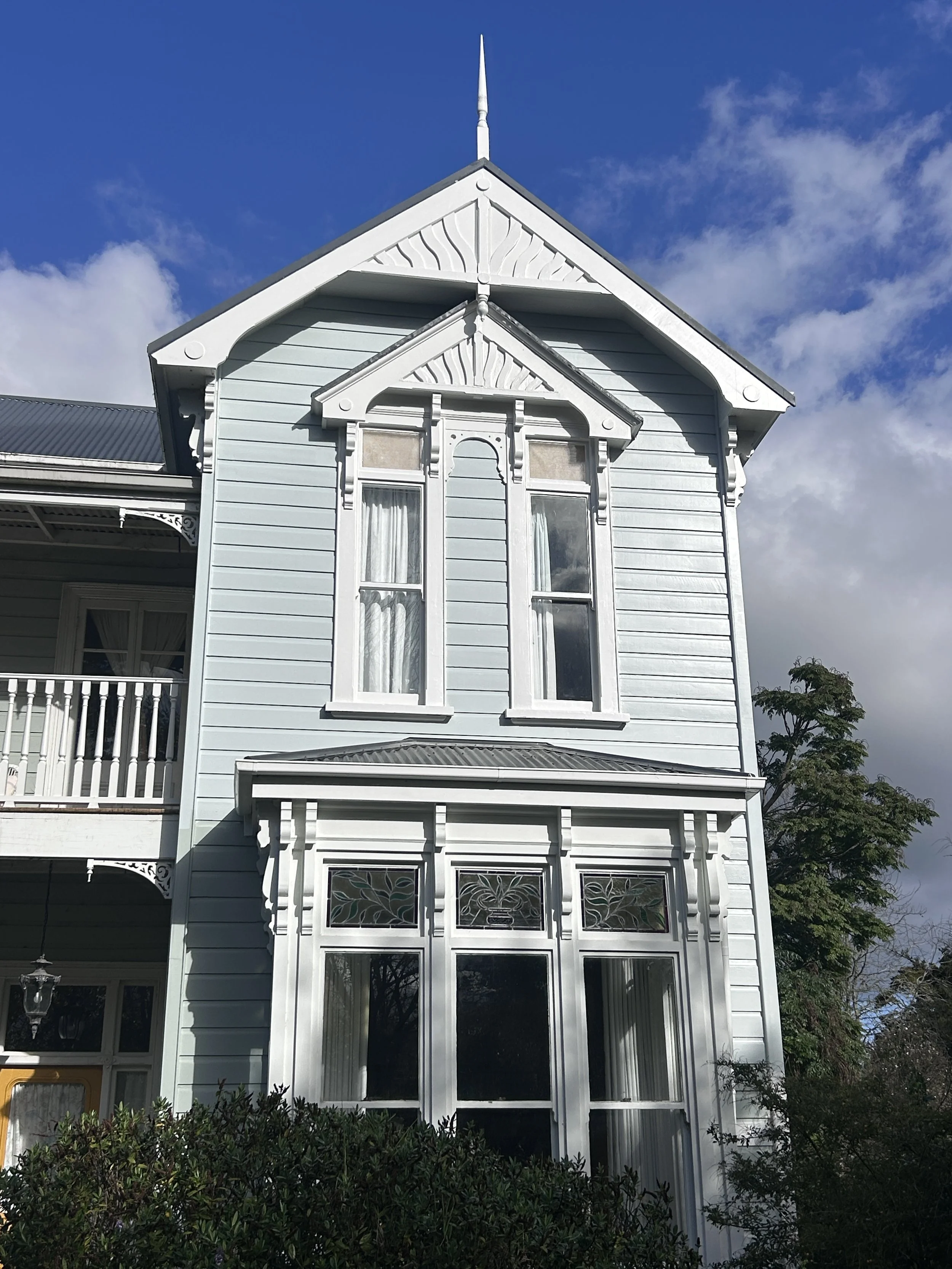 A white Victorian-style house with ornate trim, stained glass windows, and a pointed roof under a partly cloudy blue sky.