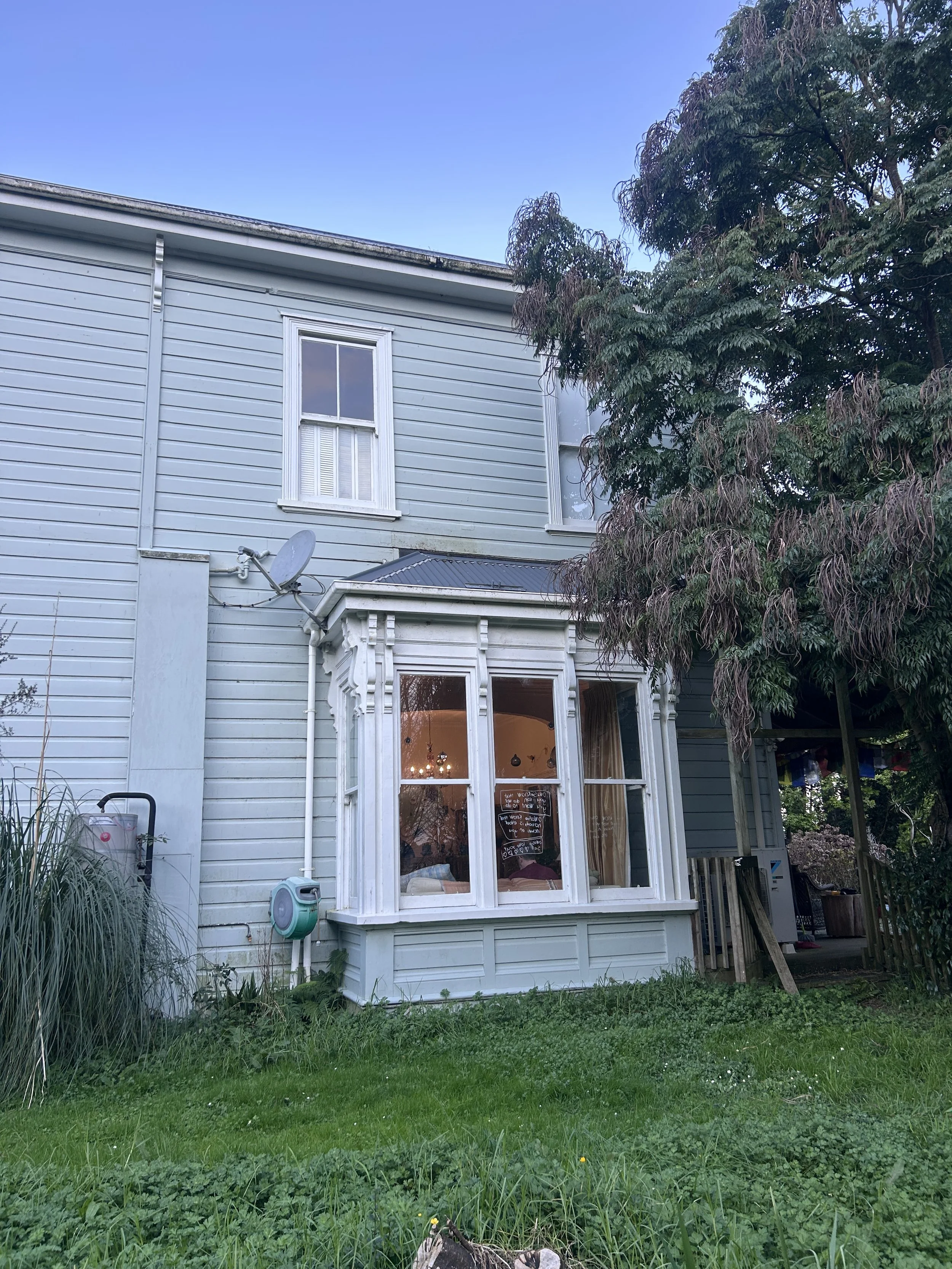 The image shows the exterior of a light blue house with white trim and siding. There are two windows on the upper level and a bay window on the lower level with curtains and some chalkboards inside. A satellite dish is attached to the house. The yard