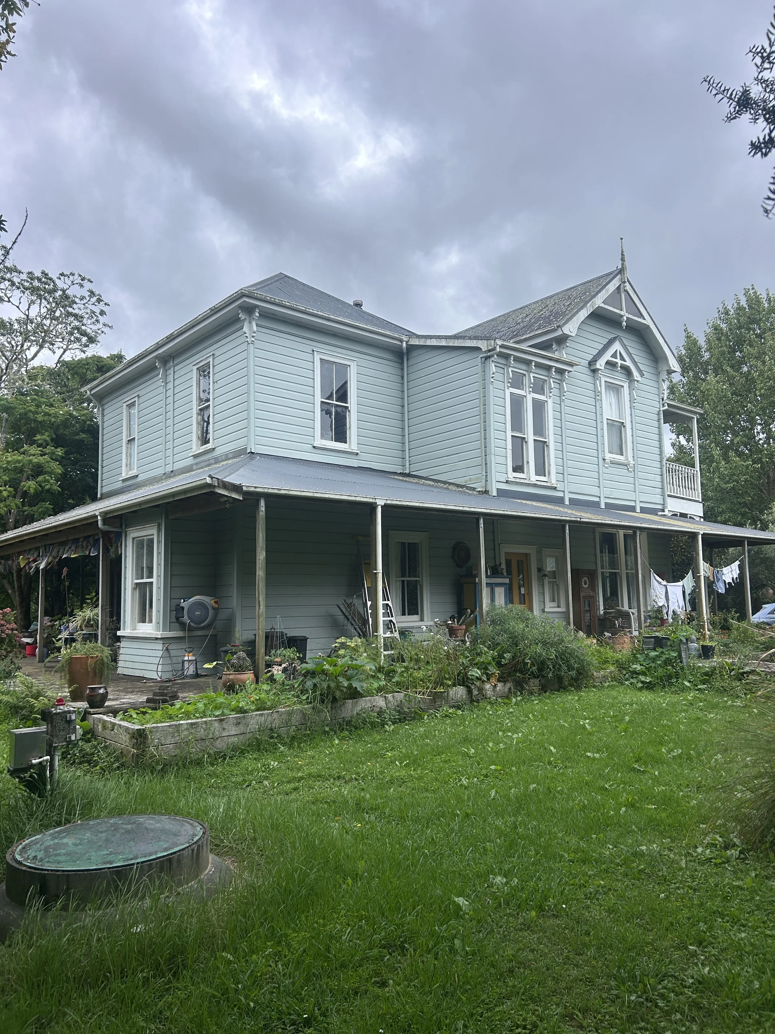 A two-story light blue house with white window frames and trim, surrounded by a green lawn and garden, with a cloudy sky overhead.