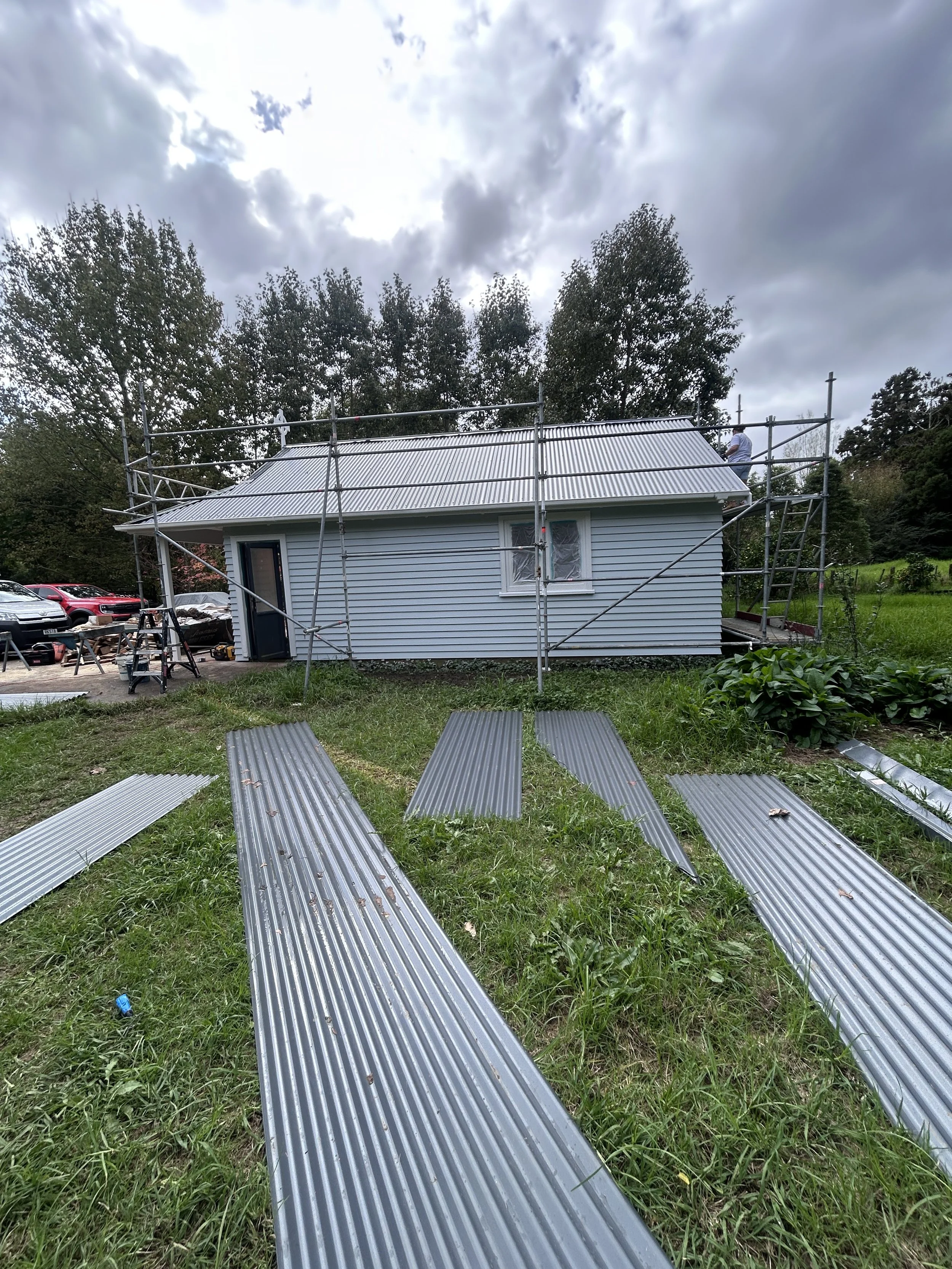A house under construction or renovation with scaffolding around it, metal roofing being installed, and various building tools and materials on the ground. Several parked cars are visible nearby, and the sky is cloudy.