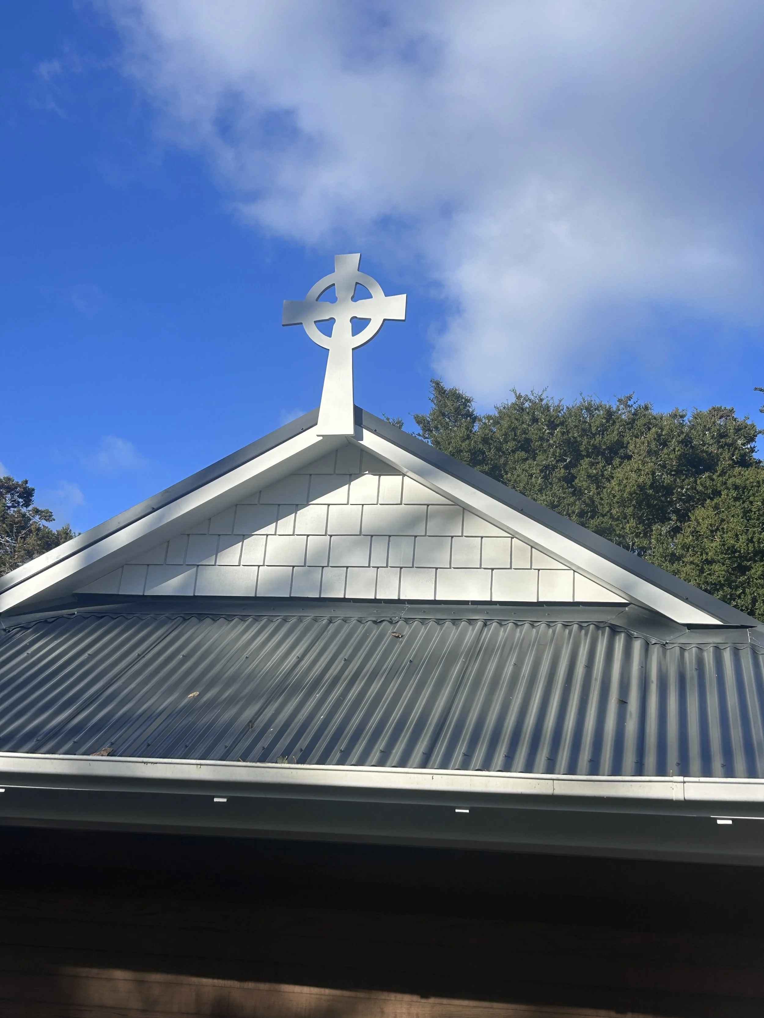 White church steeple with a cross at the top, against a partly cloudy blue sky, trees in the background, and a metal roof in the foreground.