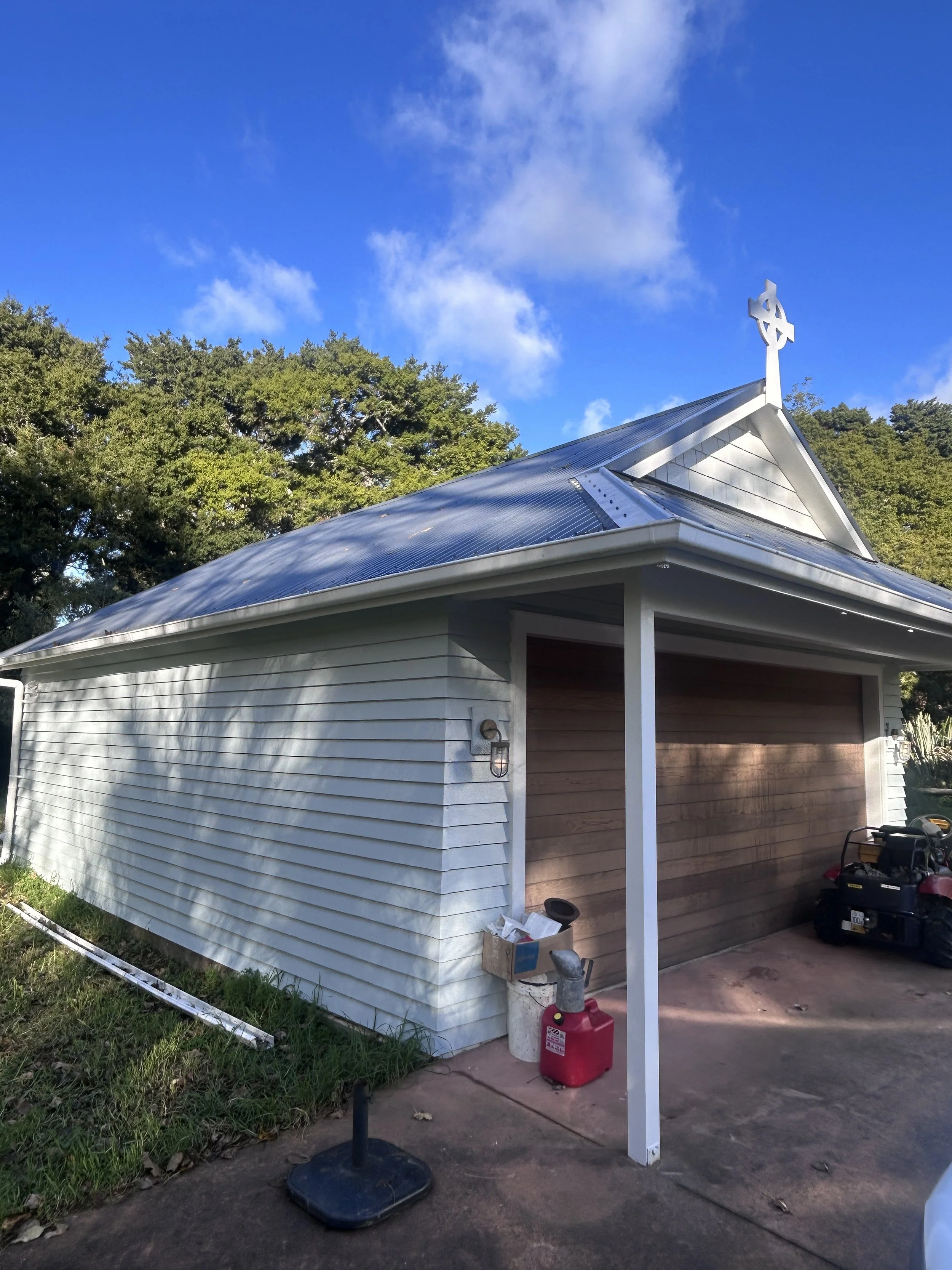 A small building with white siding and a wooden garage door. The roof is metal with a cross at the peak. There are tools and equipment outside, including a red fuel container, a small black lawn mower, and a cardboard box. The building is next to gre