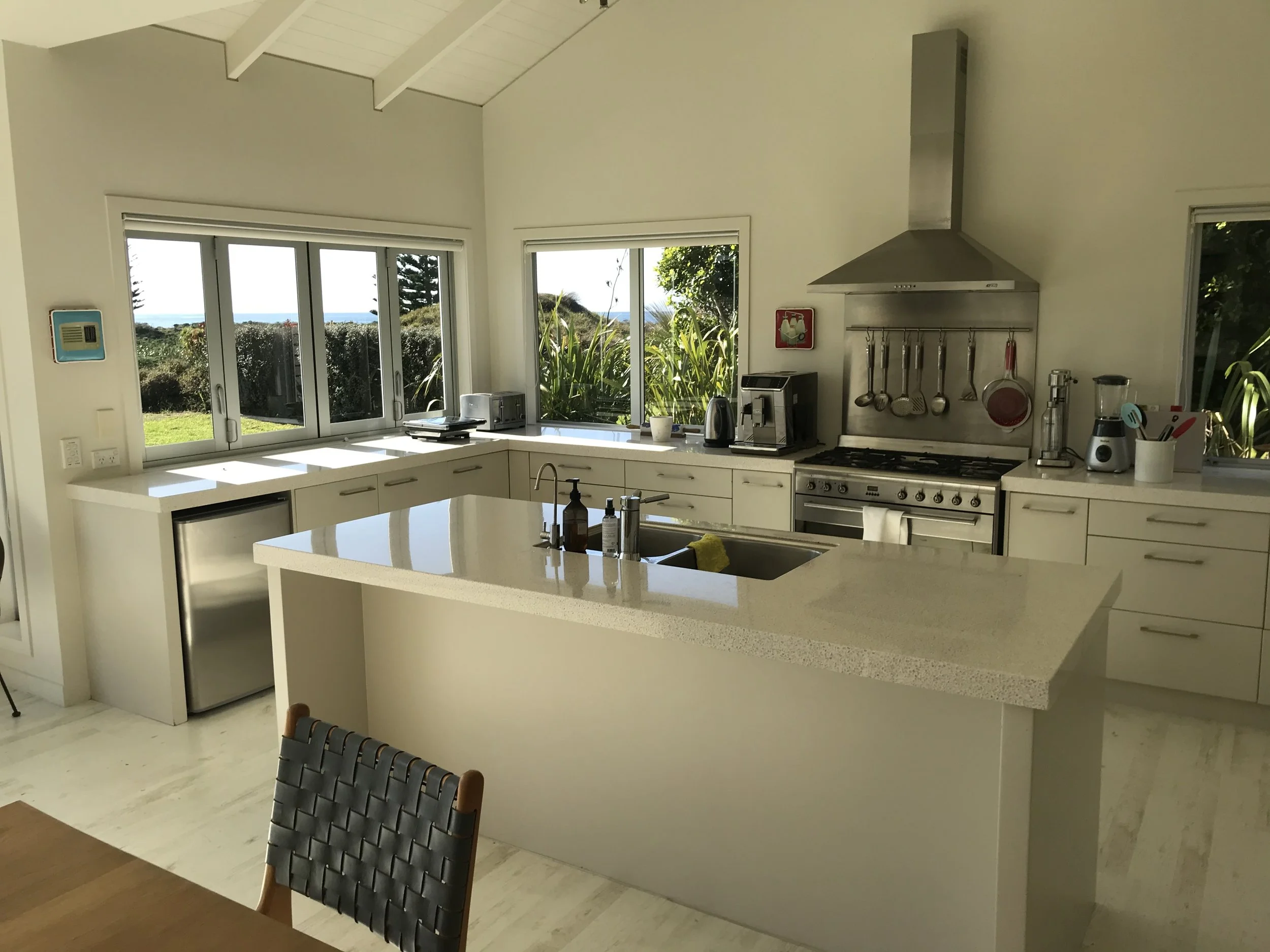 Modern kitchen with white cabinets, stainless steel appliances, large windows showing outdoors with greenery and trees, and a dark wicker chair at a wooden table.