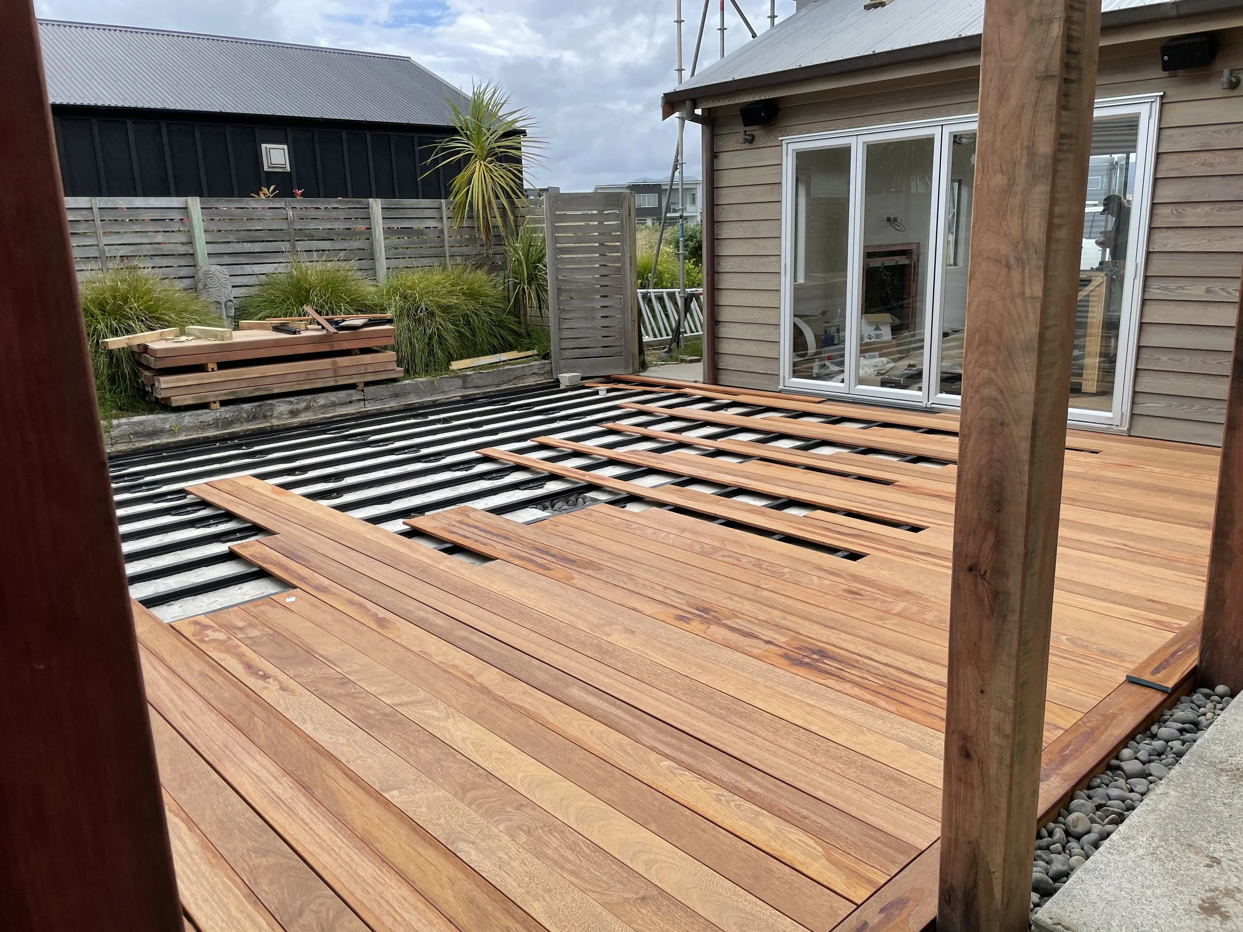 A wooden deck under construction with reddish-brown wooden planks laid out, a stack of additional wooden planks, and black and silver construction materials in the background. A sliding glass door leads into a house, and there is a wooden post to the