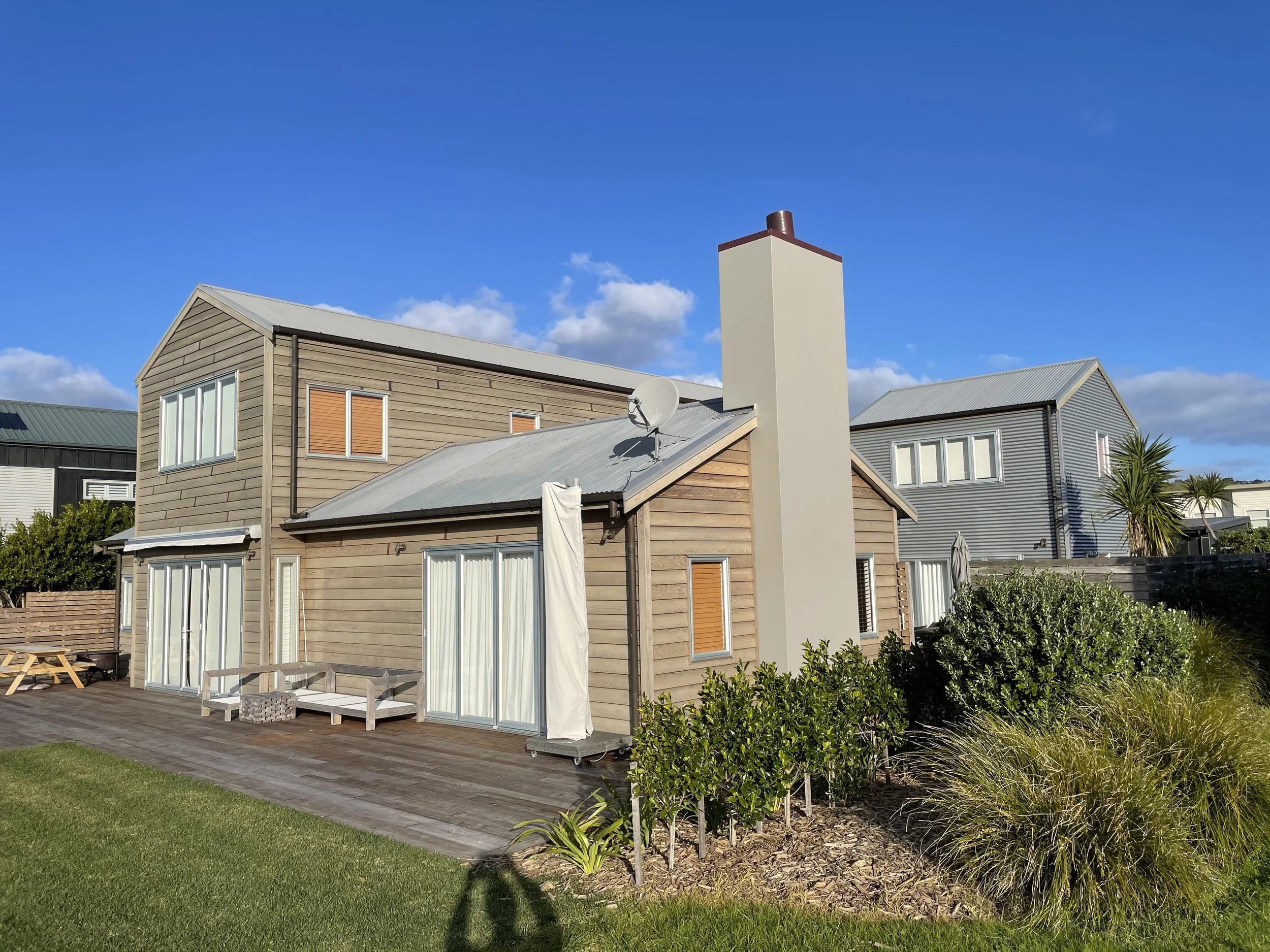 A modern residential house with a wooden exterior, large windows, and a sloped metal roof. There is a chimney, a satellite dish, and a patio area with outdoor furniture. The yard has green grass, shrubs, and small trees, all under a bright blue sky w