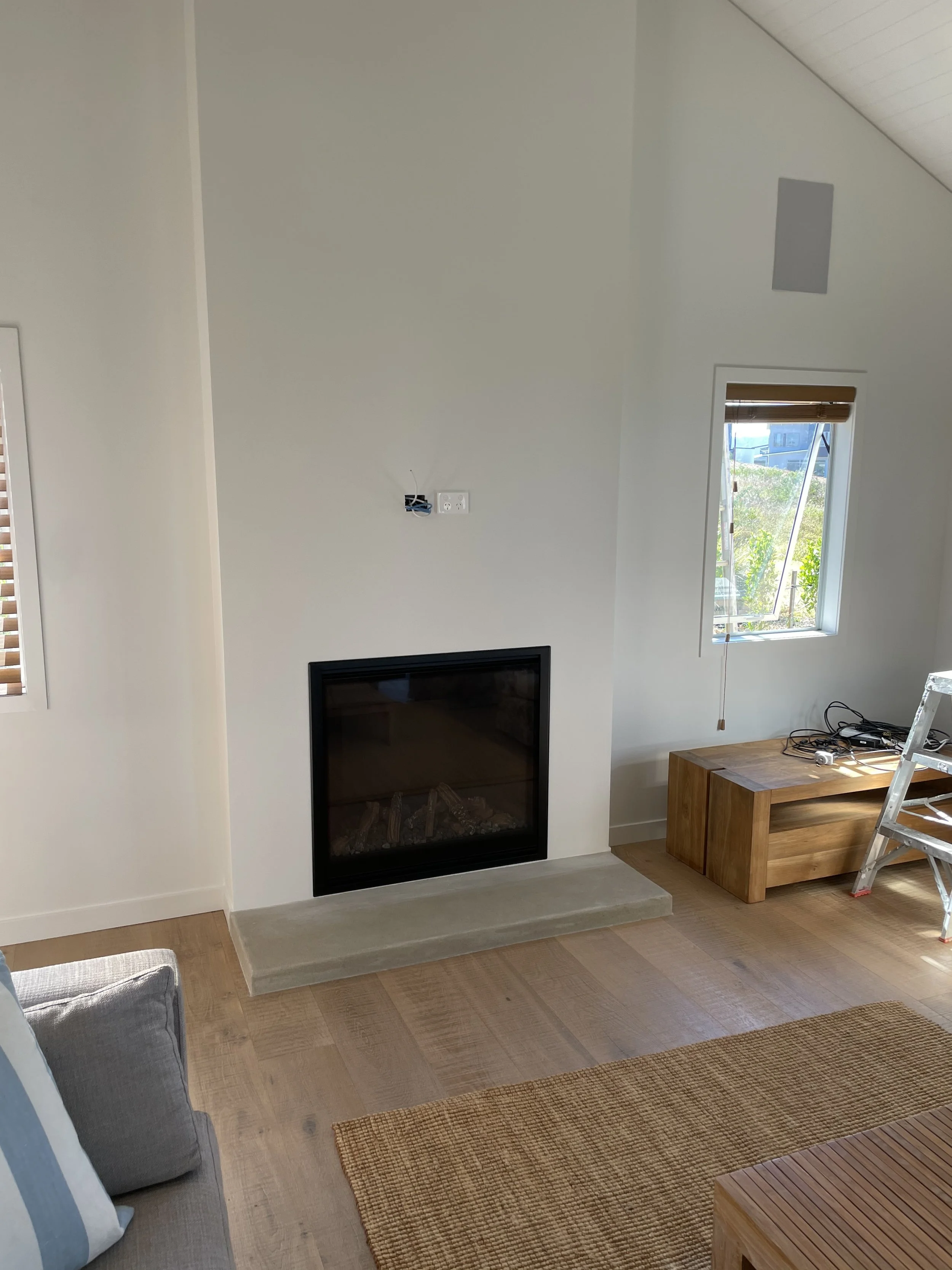 Living room with a fireplace, a wooden coffee table, and a window showing greenery outside, with part of a gray sofa visible in the foreground.