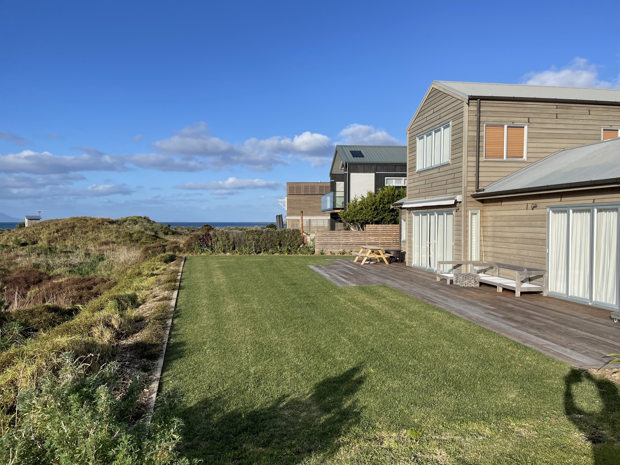 View of modern houses with large windows, a wooden deck, and green lawn in a coastal area under blue sky with clouds.