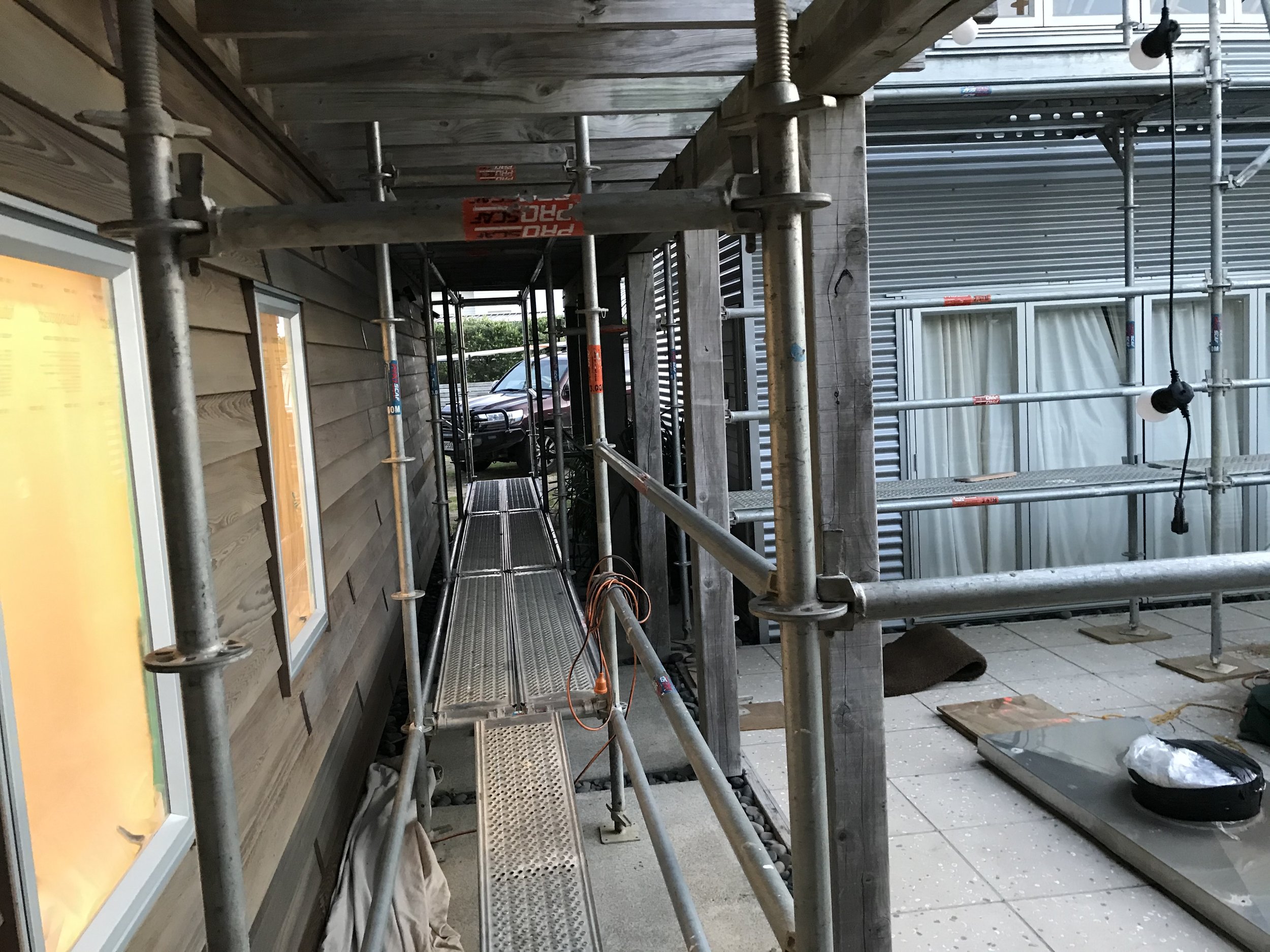 Construction scaffolding set up along the exterior wall of a building, with some construction materials and tools on the ground nearby.