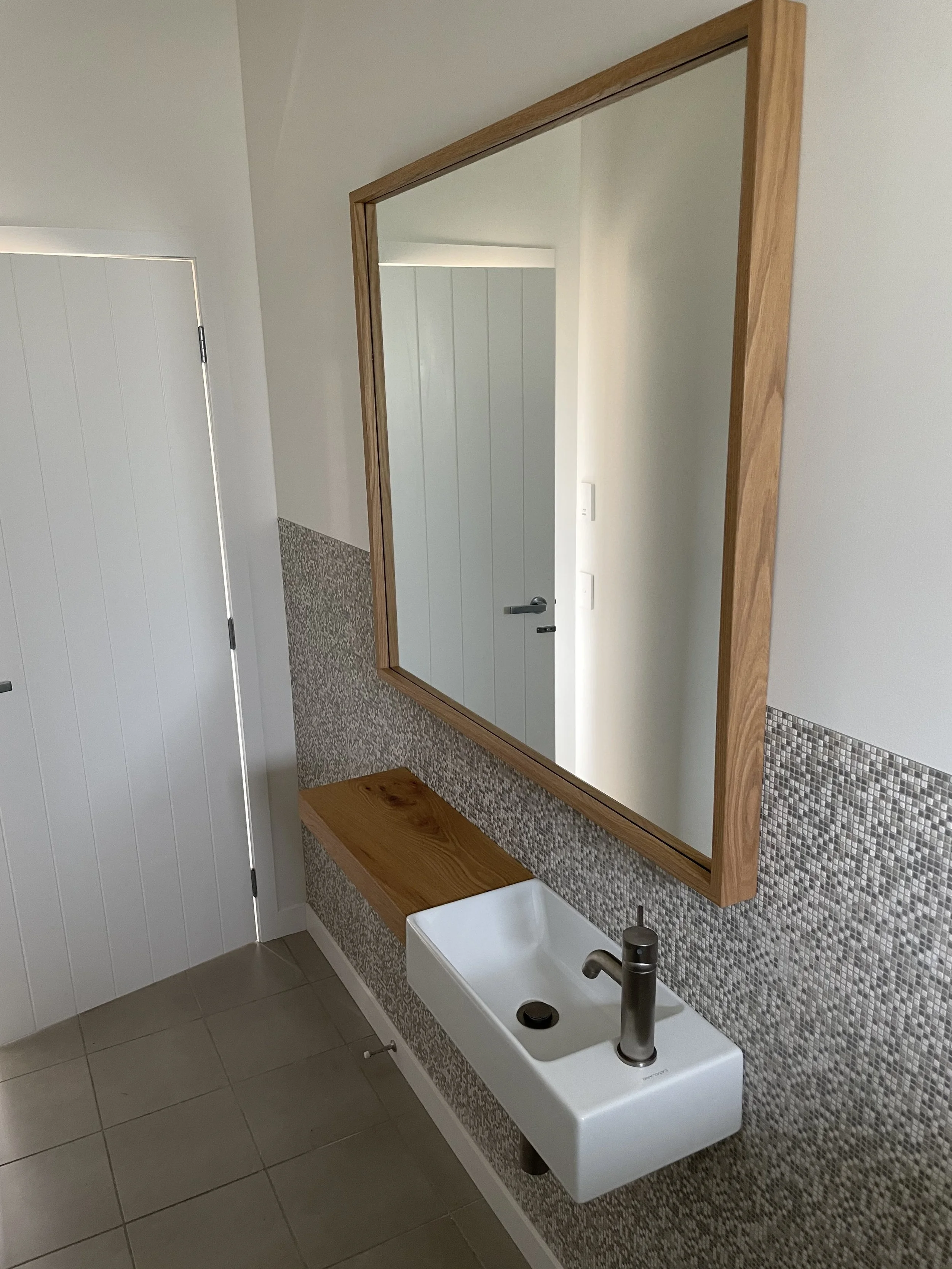 A modern bathroom with a rectangular mirror framed in wood, mounted above a white sink with a metallic faucet. The wall behind the mirror has a mosaic tile accent strip, and the sink is attached beneath a wooden shelf. To the left is a white paneled 