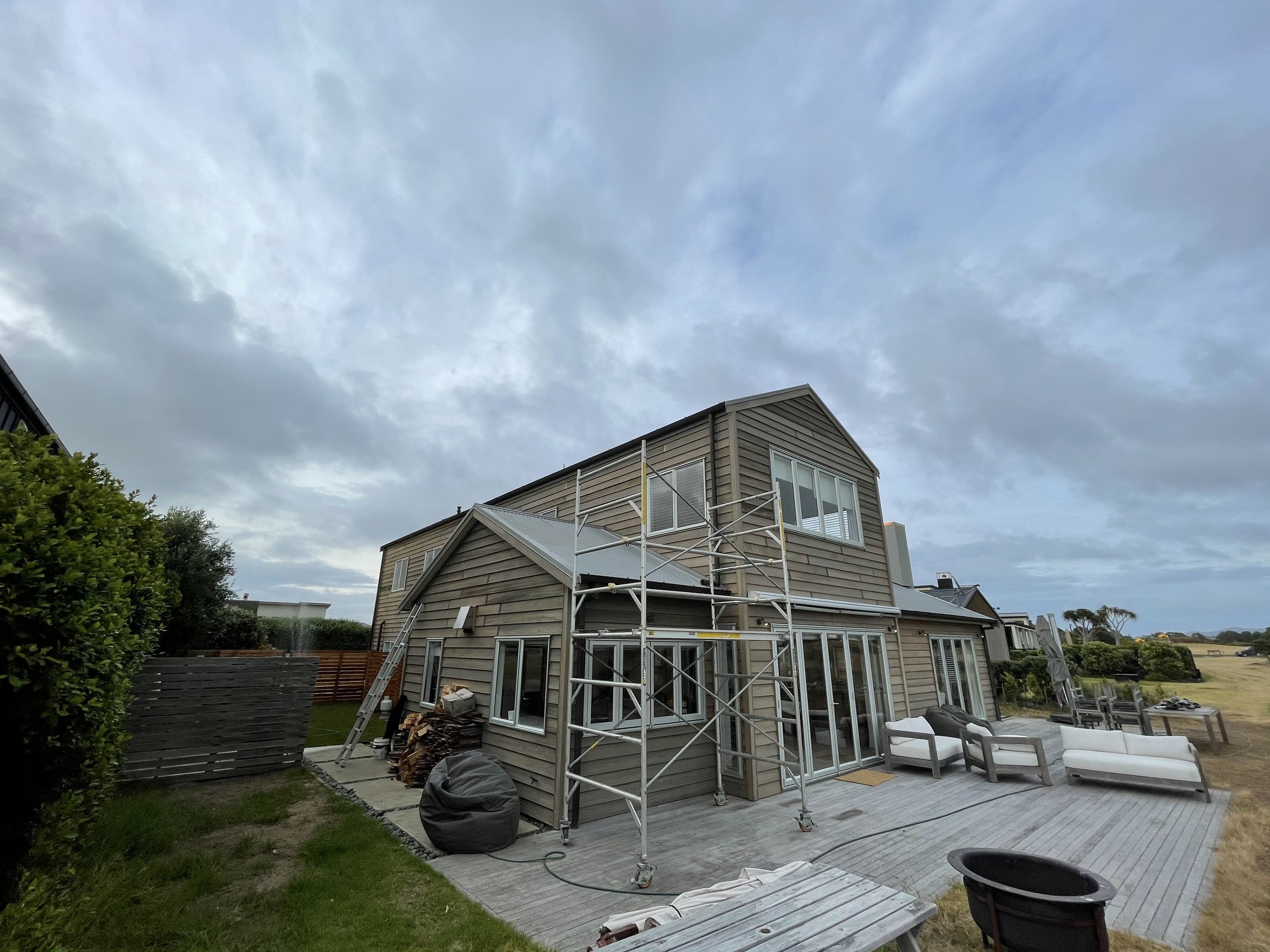 A two-story house with construction scaffolding around it, located in a backyard with a wooden deck, outdoor furniture, and a lawn, under a cloudy sky.