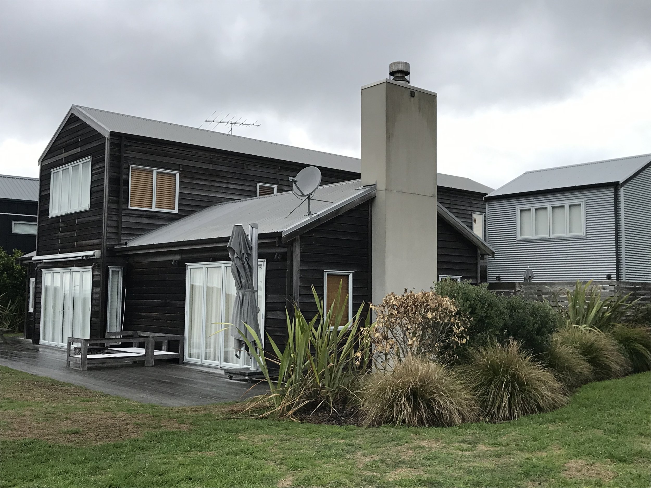 A modern house with black wooden exterior walls, white window frames, and a metal roof. There is a satellite dish attached to the roof and a chimney on top. The house has sliding glass doors opening to a wooden deck, with patio furniture and an umbre