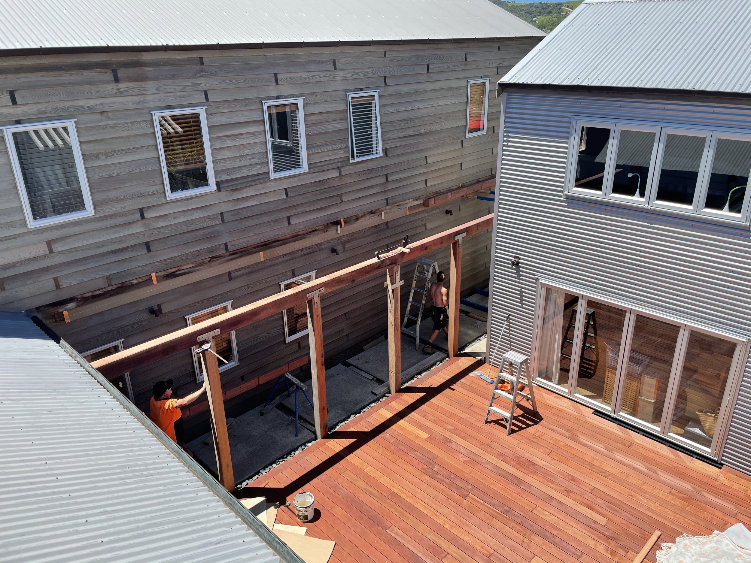 Two houses are under construction, with workers installing a wooden deck between them. One house has beige brick siding and the other has metal siding.