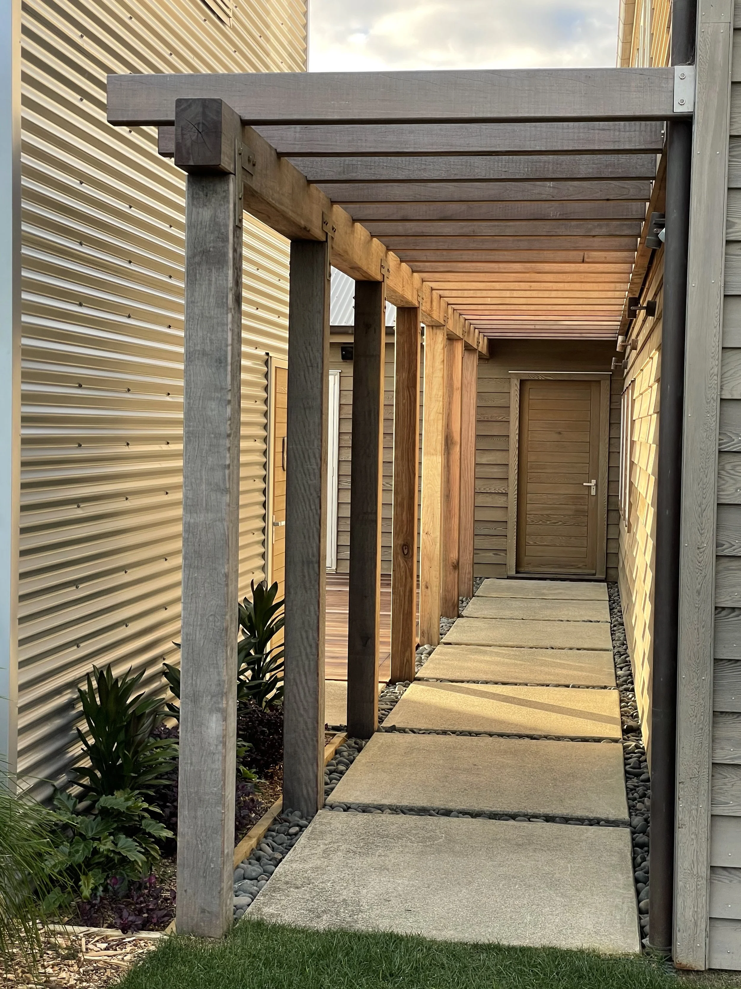 A wooden pathway with square stepping stones leads to a door in a backyard. The pathway is framed by a wooden frame on one side and is adjacent to a beige metal wall on the other. There are plants with large green leaves on the left side of the pathw