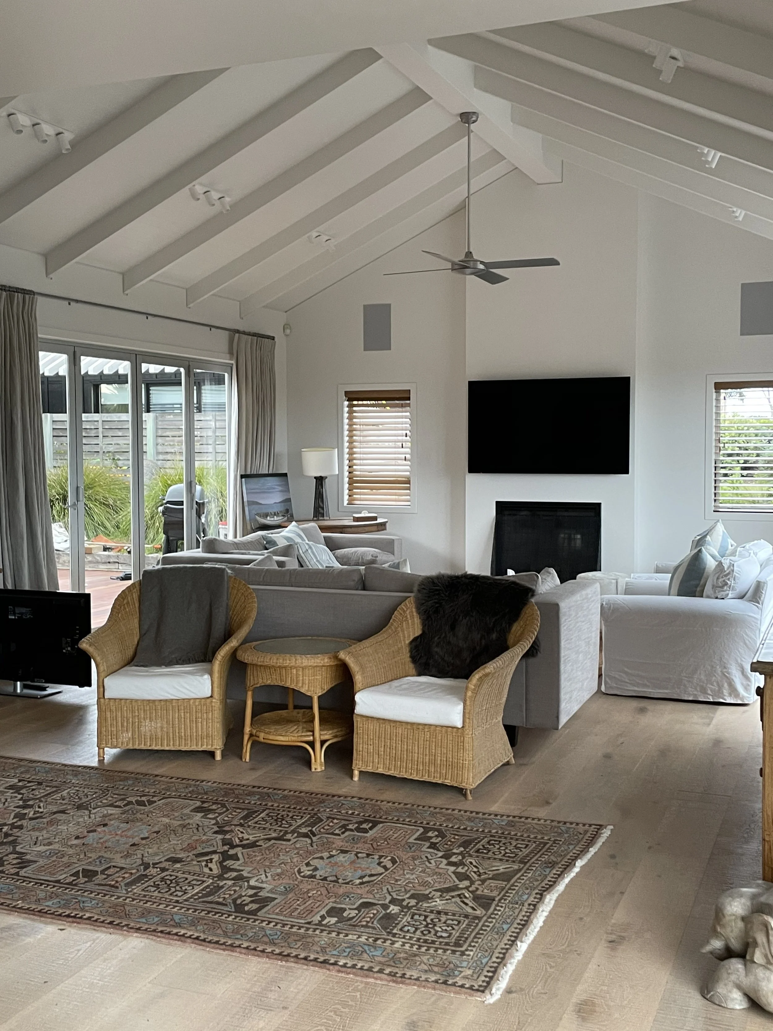 Living room with beige carpet, ornate rug, white sofas, wicker chairs, large window with wooden blinds, sliding glass door to patio, black TV on wall above fireplace, ceiling fan, and large windows with curtains.