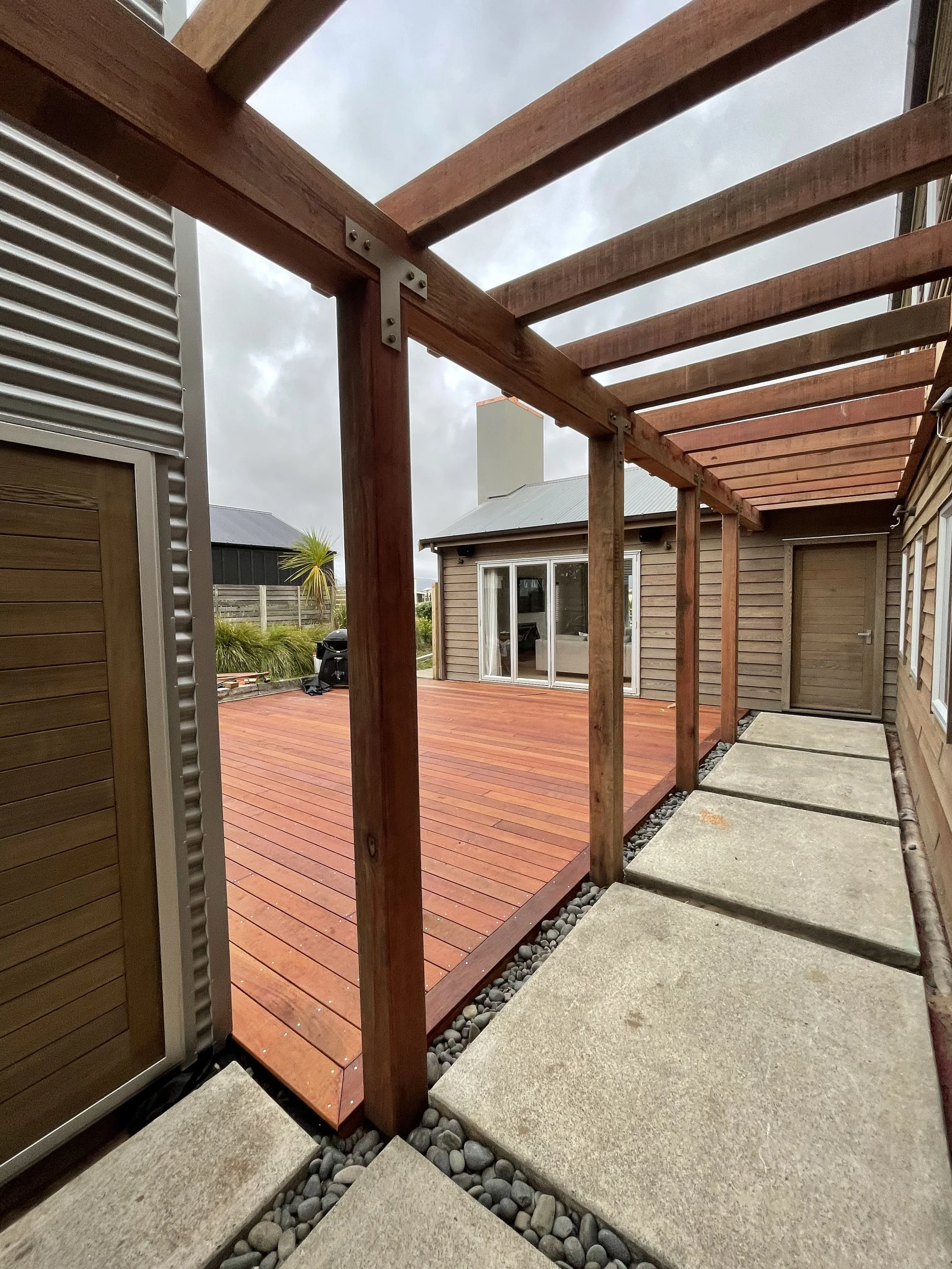 A view of a wooden deck under construction with a wooden pergola frame, concrete stepping stones on the side, and a neighboring house in the background.
