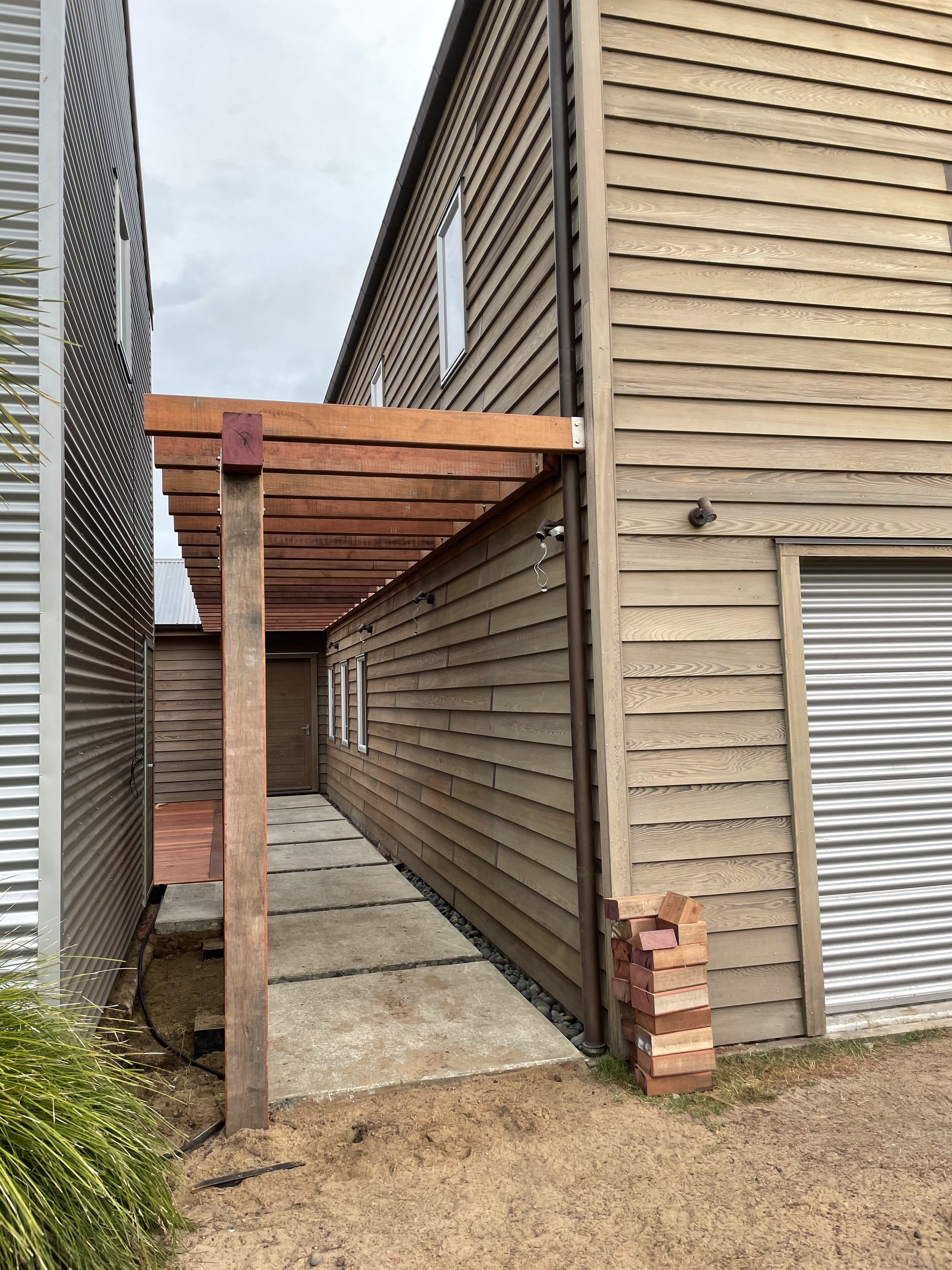 Image showing the side of a modern wooden house with horizontal siding, windows, and a partially constructed wooden pergola, near a metal wall with horizontal siding and some construction materials on the ground.