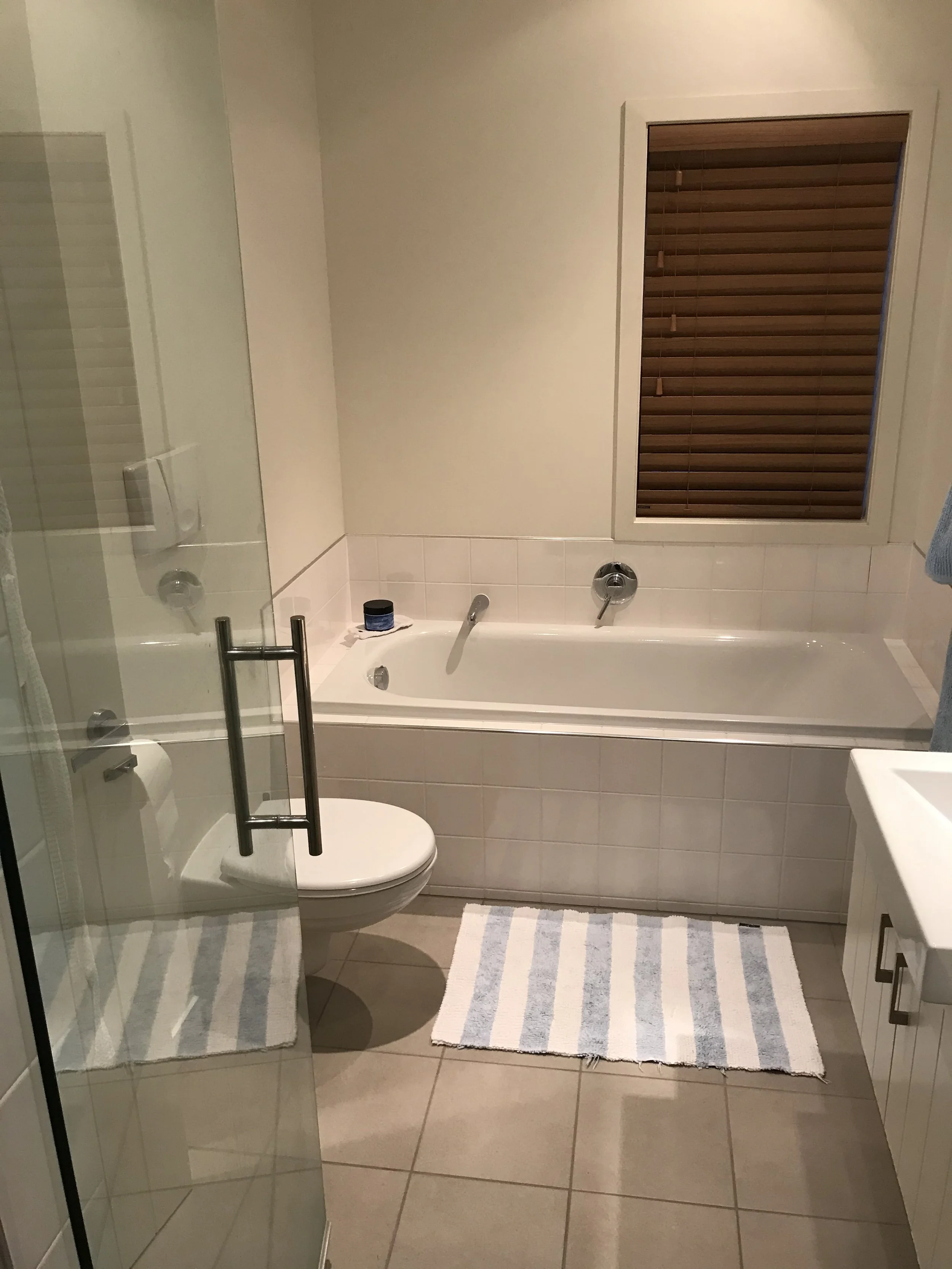 A bathroom with a bathtub, a toilet, a striped bath mat, and a window with brown blinds. The bathroom has beige tiled floors and white tiled walls.