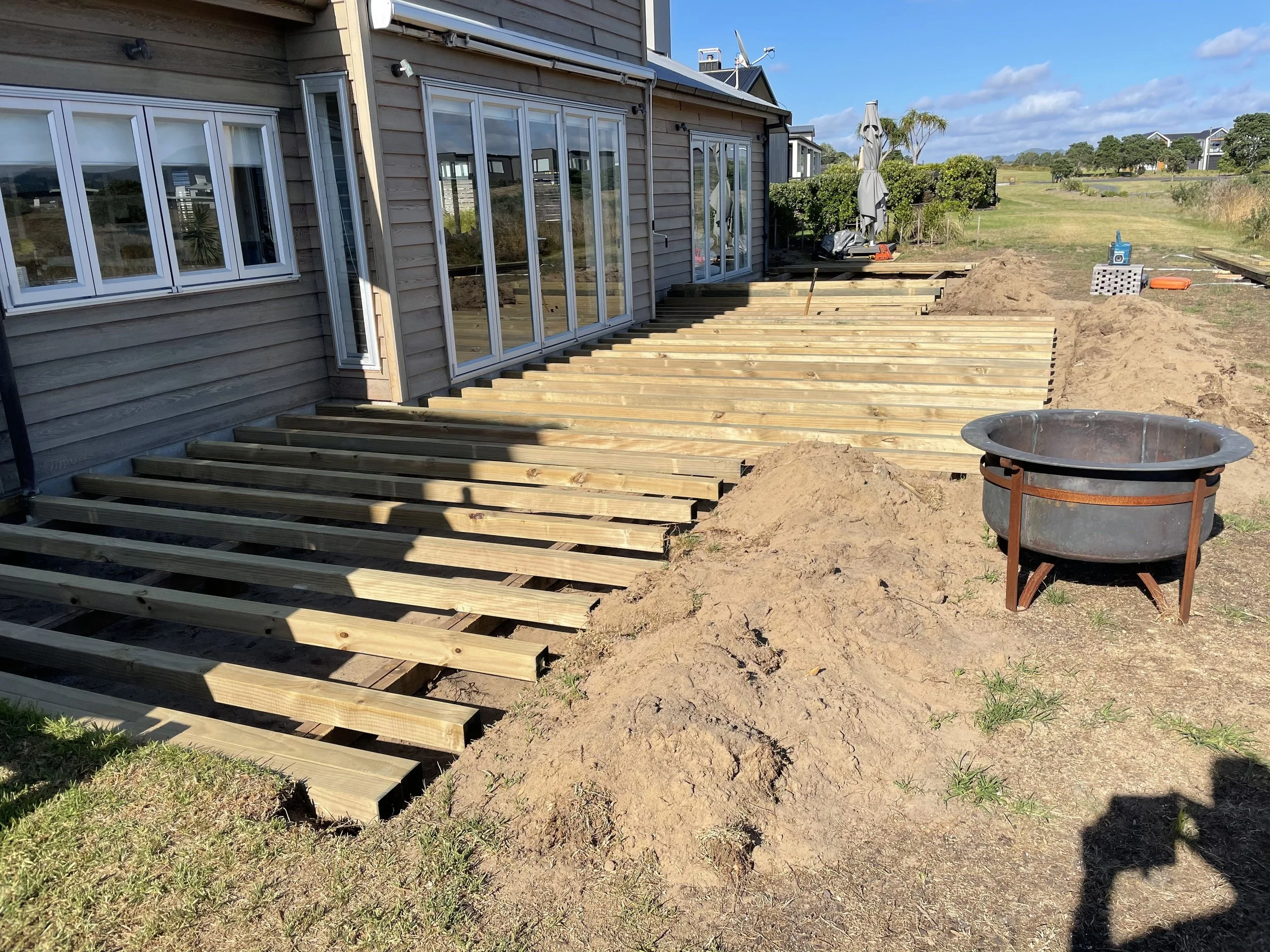 A house with large sliding glass doors, undergoing construction with a new deck being built. Wooden beams are laid out, and there are tools and materials on site including an umbrella, a speaker, and construction debris. The background shows a grassy