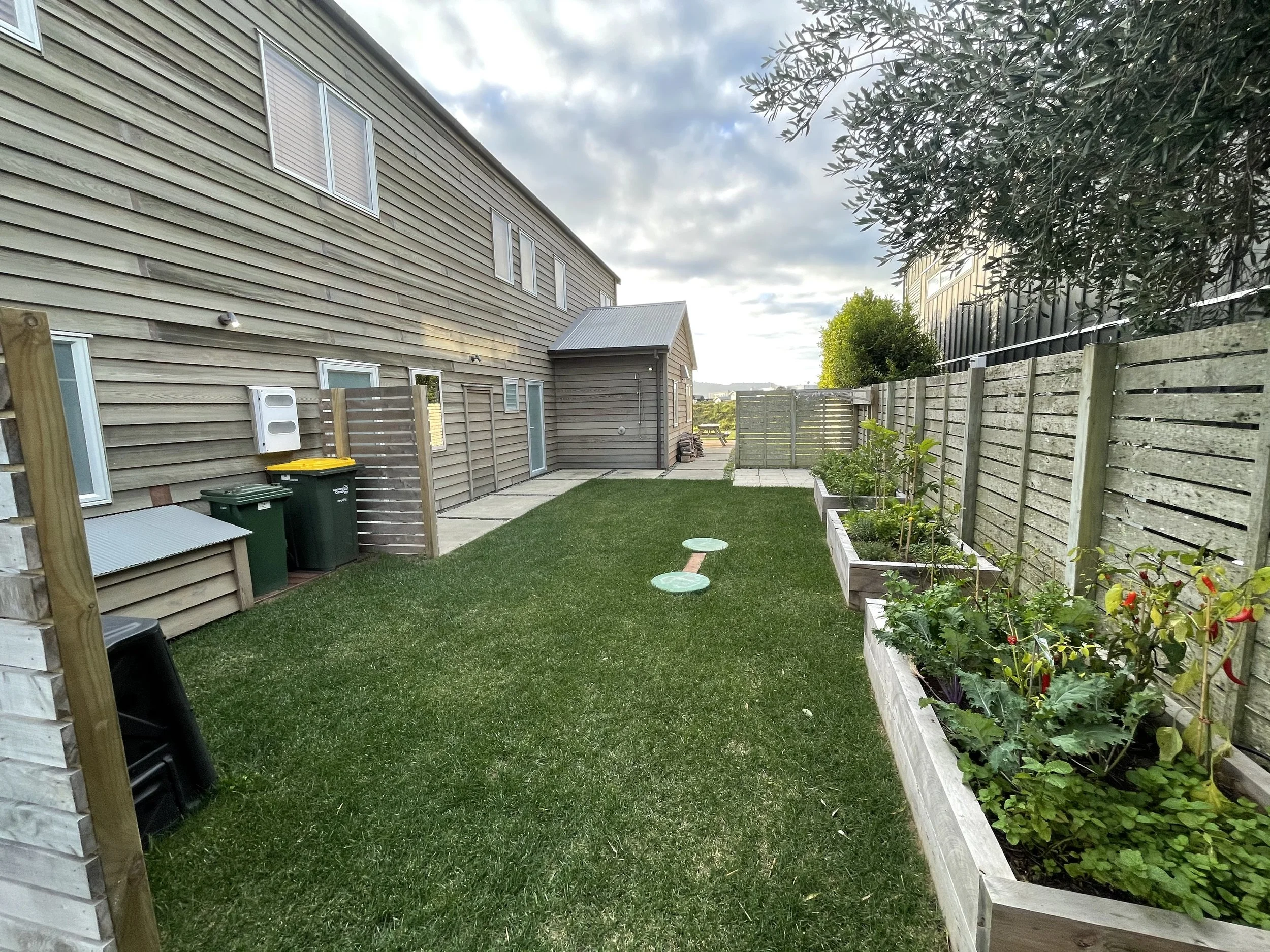 A backyard with a brick and wood house, a lawn, raised garden beds with plants, a paved pathway, a small shed, and a wooden fence with trees in the background.