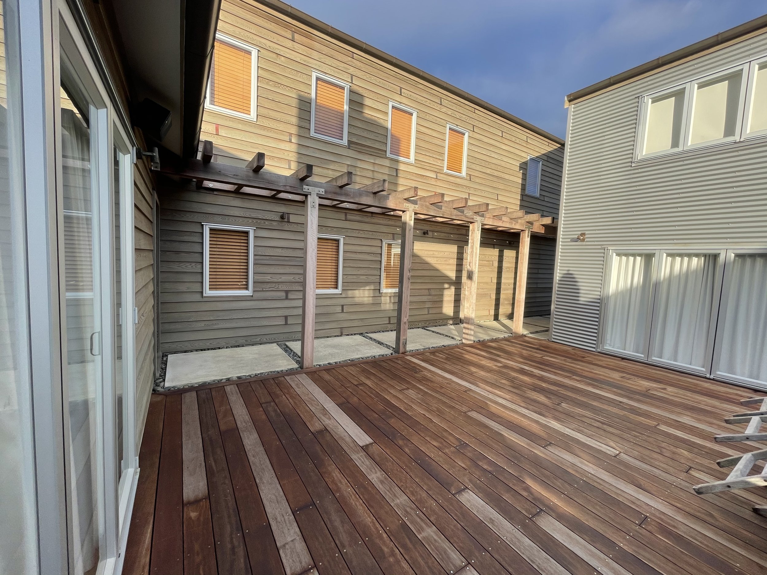 A spacious outdoor wooden deck connected to a modern house with large windows and sliding glass doors. The house has light-colored siding, and there is a small pergola frame built on the deck, with a clear blue sky overhead.