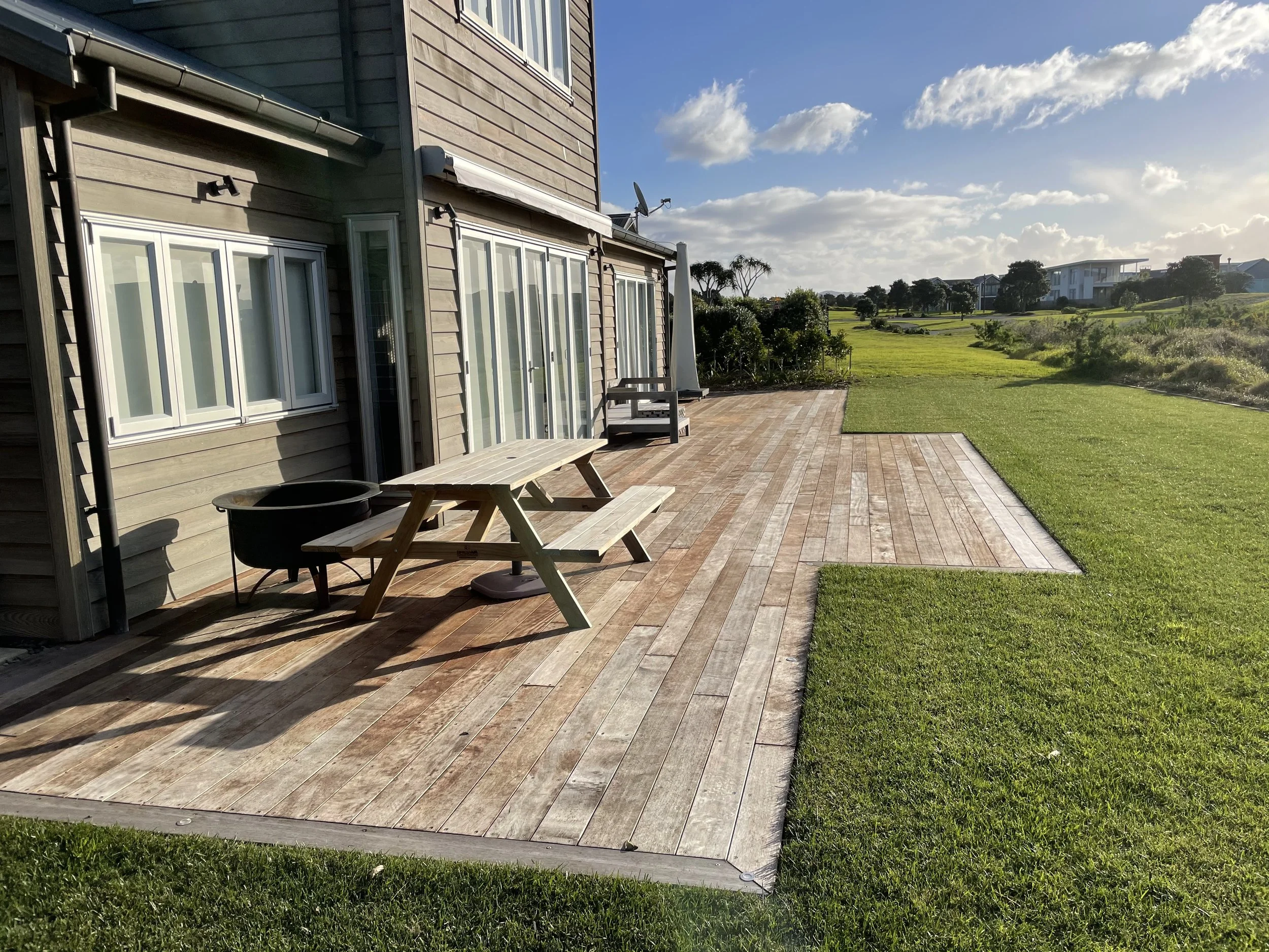 A backyard patio with a wooden deck, outdoor furniture including a picnic table and a sun lounger, and a view of a grassy lawn and distant houses under a partly cloudy sky.
