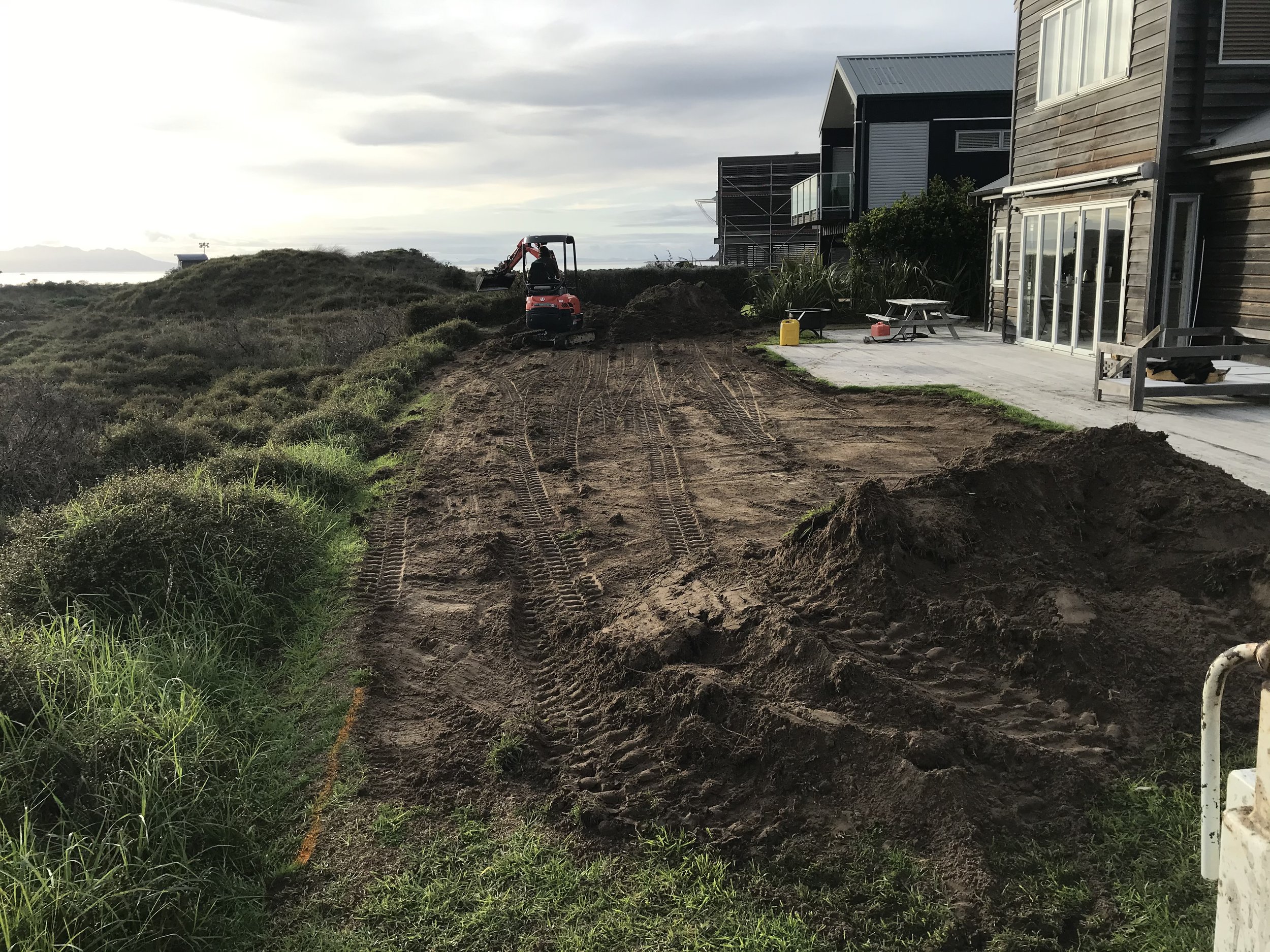 A construction site near a residential area with a small excavator digging in the dirt alongside a house with a cardboard picnic table, water containers, and other equipment, with overcast sky