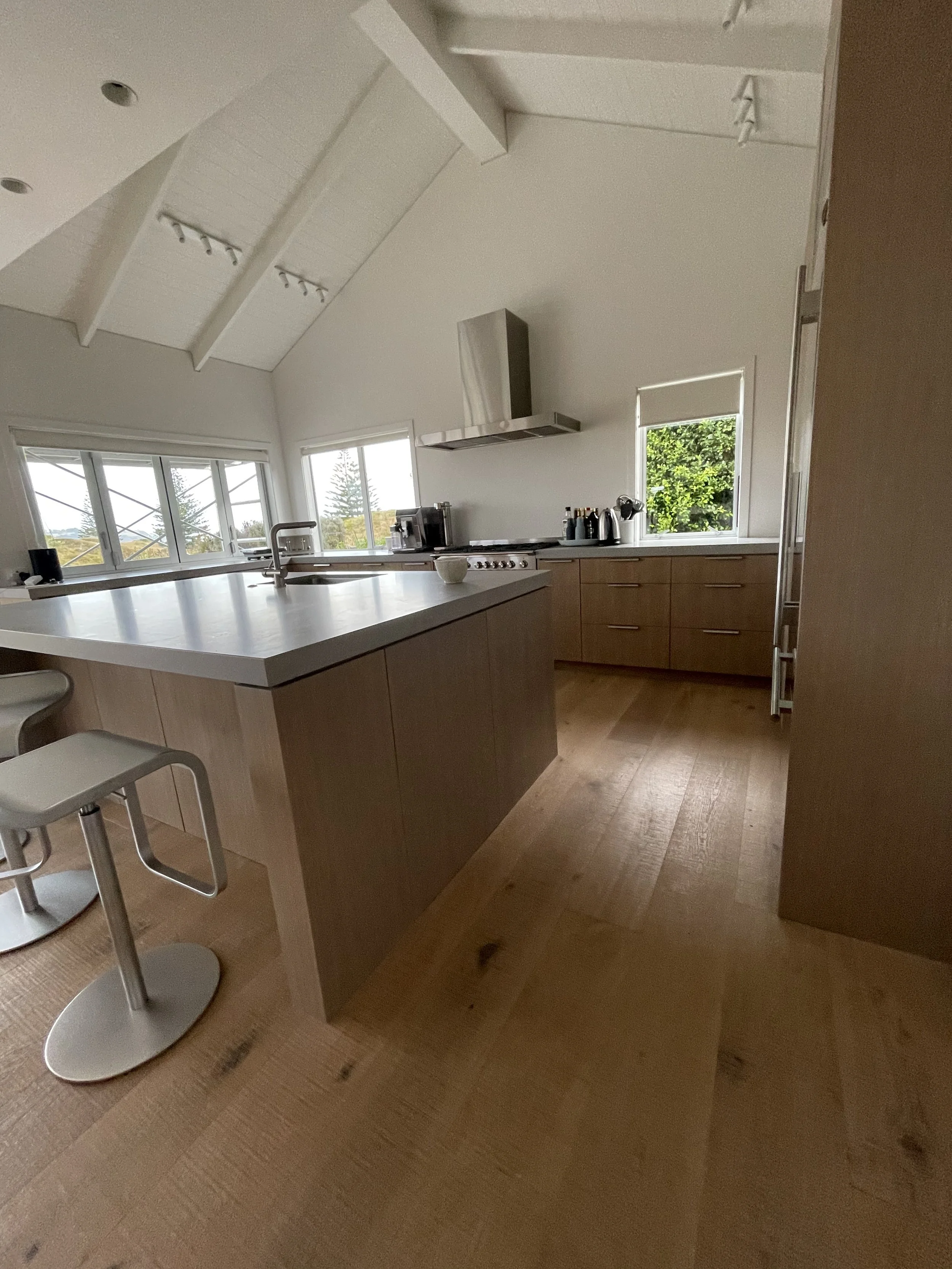 Modern kitchen with wooden cabinets, white countertop, stainless steel range hood, small window with view of greenery, kitchen appliances, and two metal bar stools.