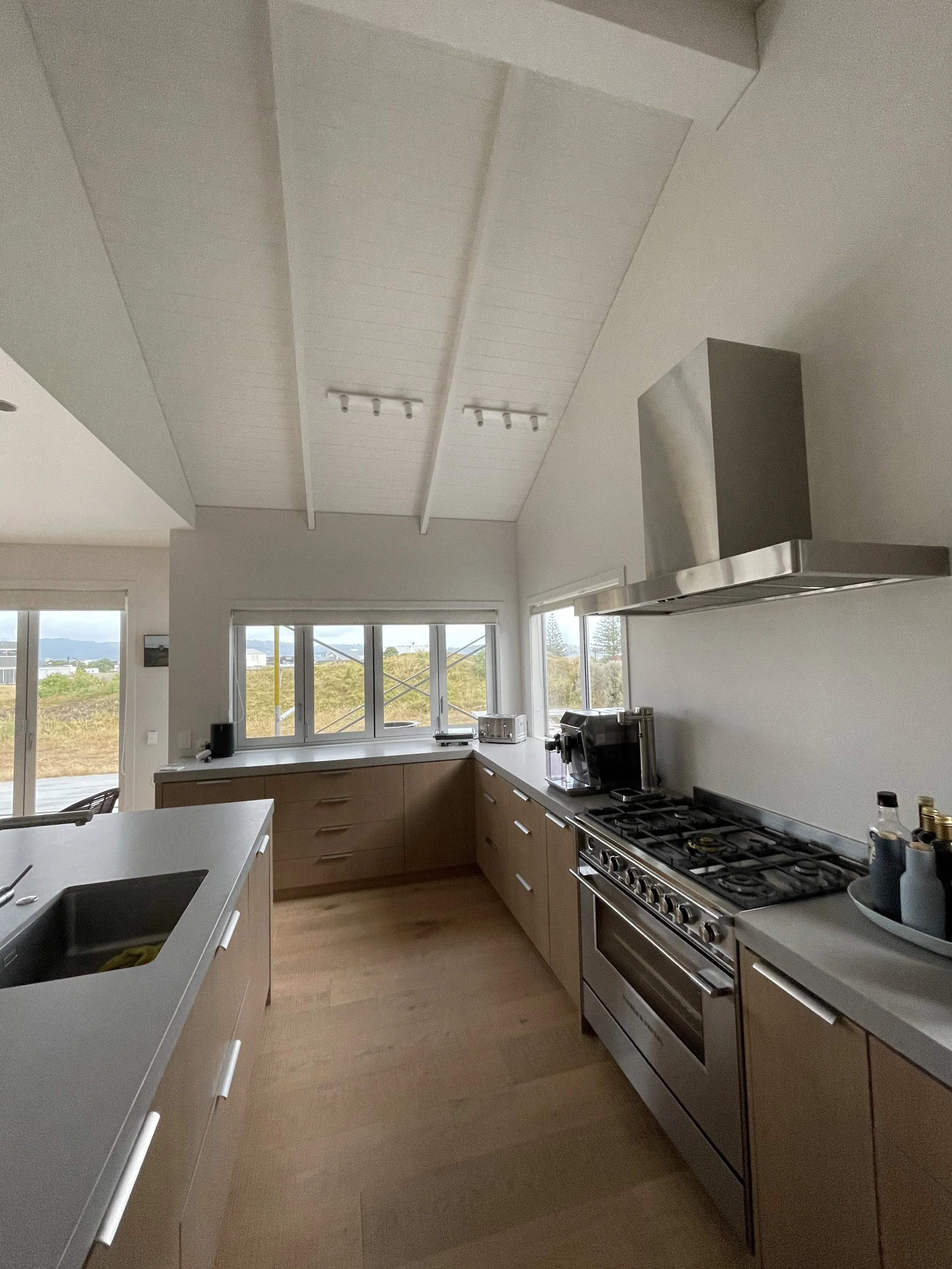Modern kitchen with wooden cabinets, stainless steel stove and range hood, white countertops, and large windows showing outdoor view.