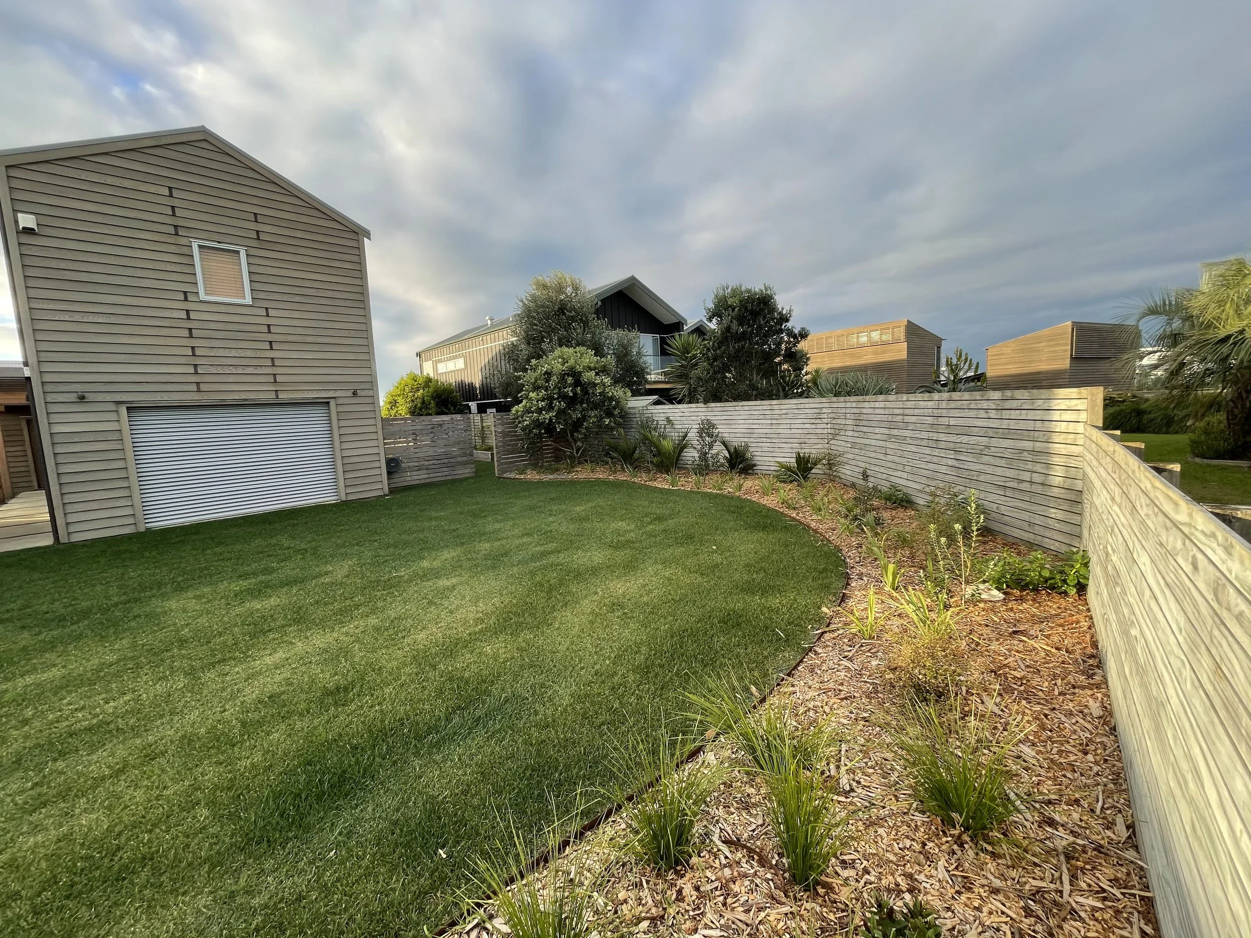 A backyard with a green lawn, a wooden fence, a few trees and bushes, and modern houses in the background under a partly cloudy sky.