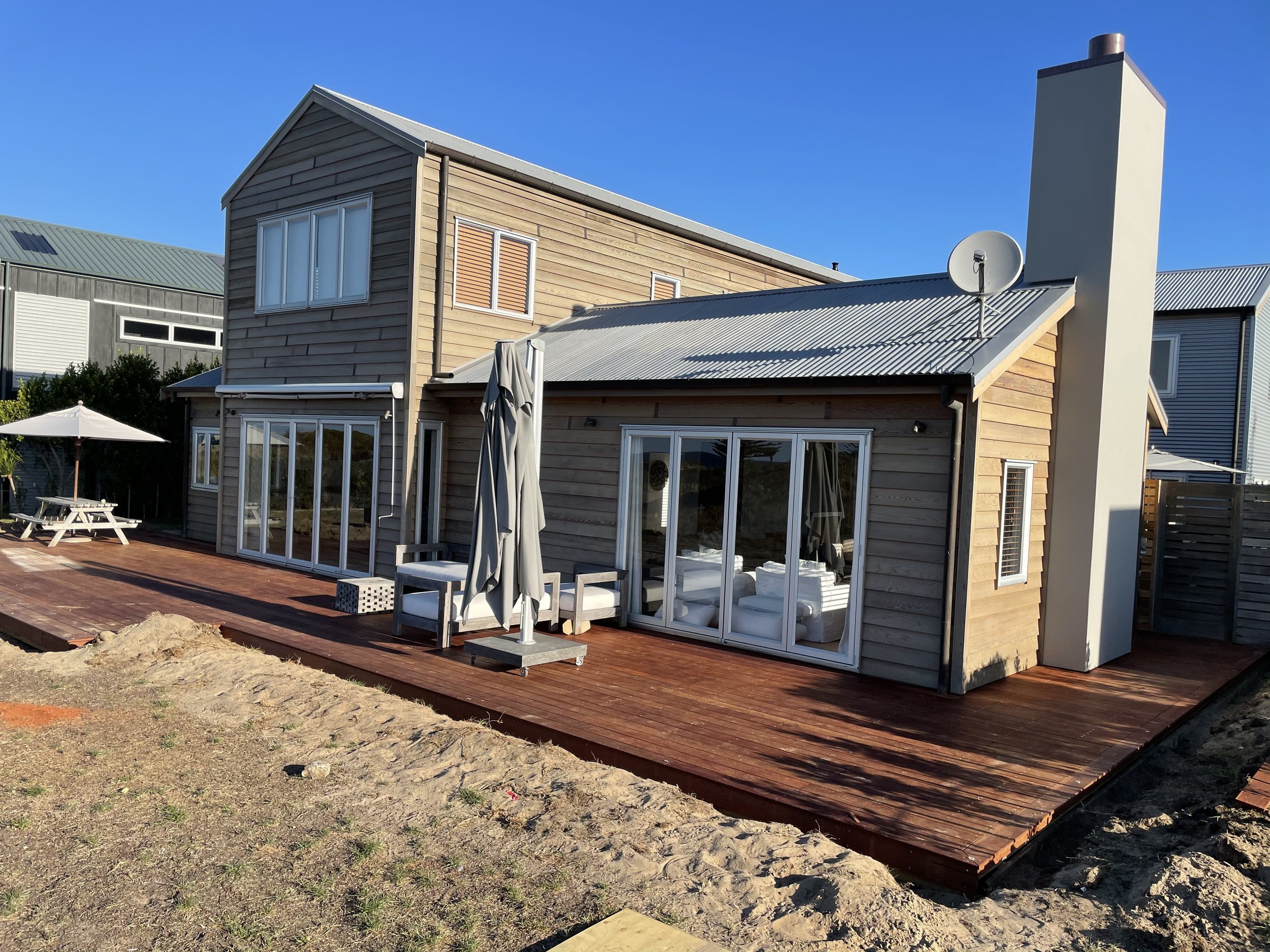 A two-story house with wooden siding, large glass sliding doors, and a metal roof. There is a satellite dish on the roof and outdoor seating with an umbrella on a wooden deck.