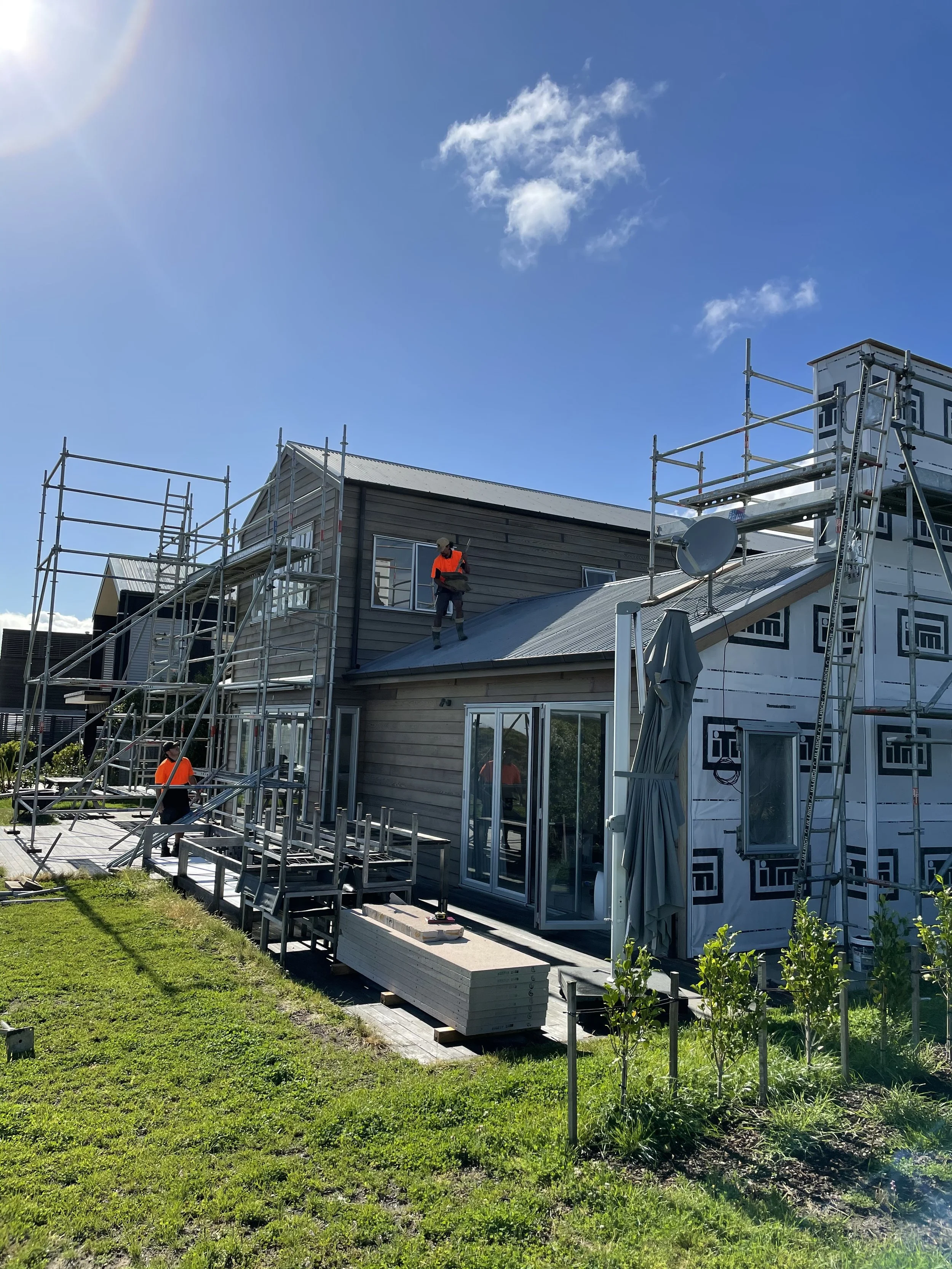 Construction workers on scaffolding working on a house with gray siding and a metal roof on a sunny day, with a lawn and plants in the foreground.