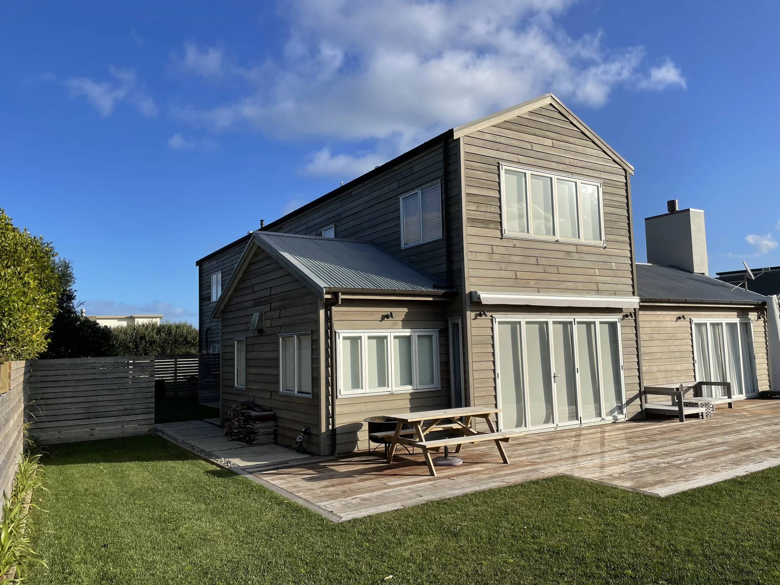 A modern house with beige wooden siding and large windows, a small lawn, and a wooden deck with a picnic table outdoors. The sky is partly cloudy with a blue background.