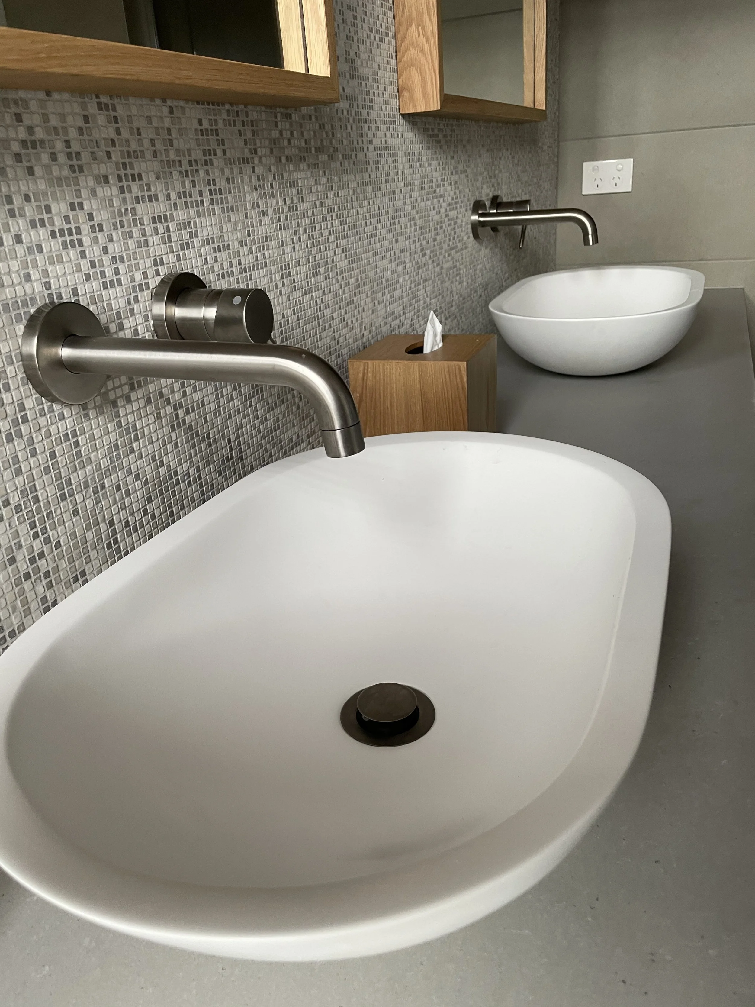 Bathroom with two modern white sinks, faucet, tissue box, tile backsplash, and electrical outlet.