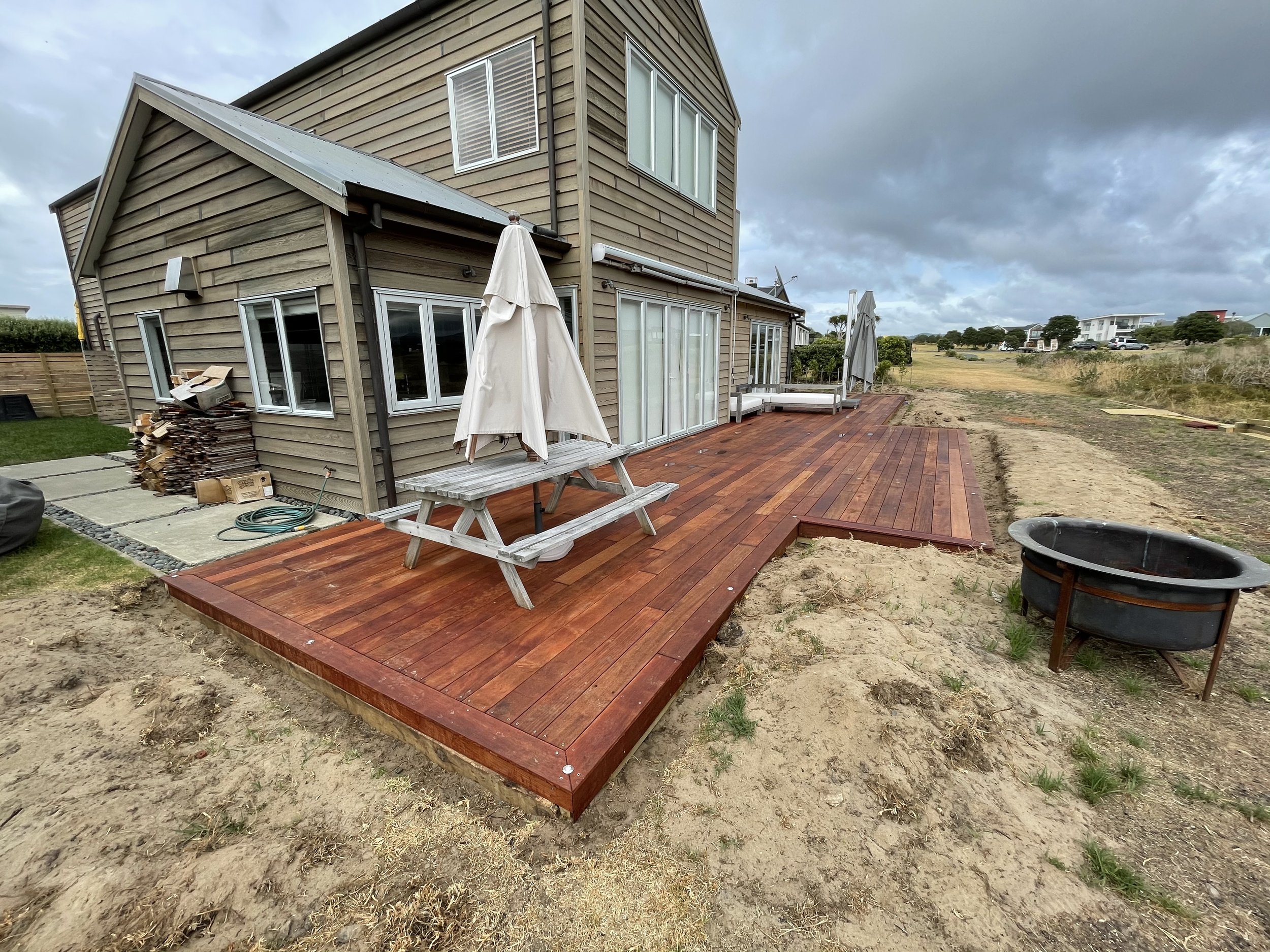A two-story house with beige wooden siding and large windows, a deck with reddish-brown wooden planks, an outdoor umbrella, and some patio furniture, with a cloudy sky and open land in the background.