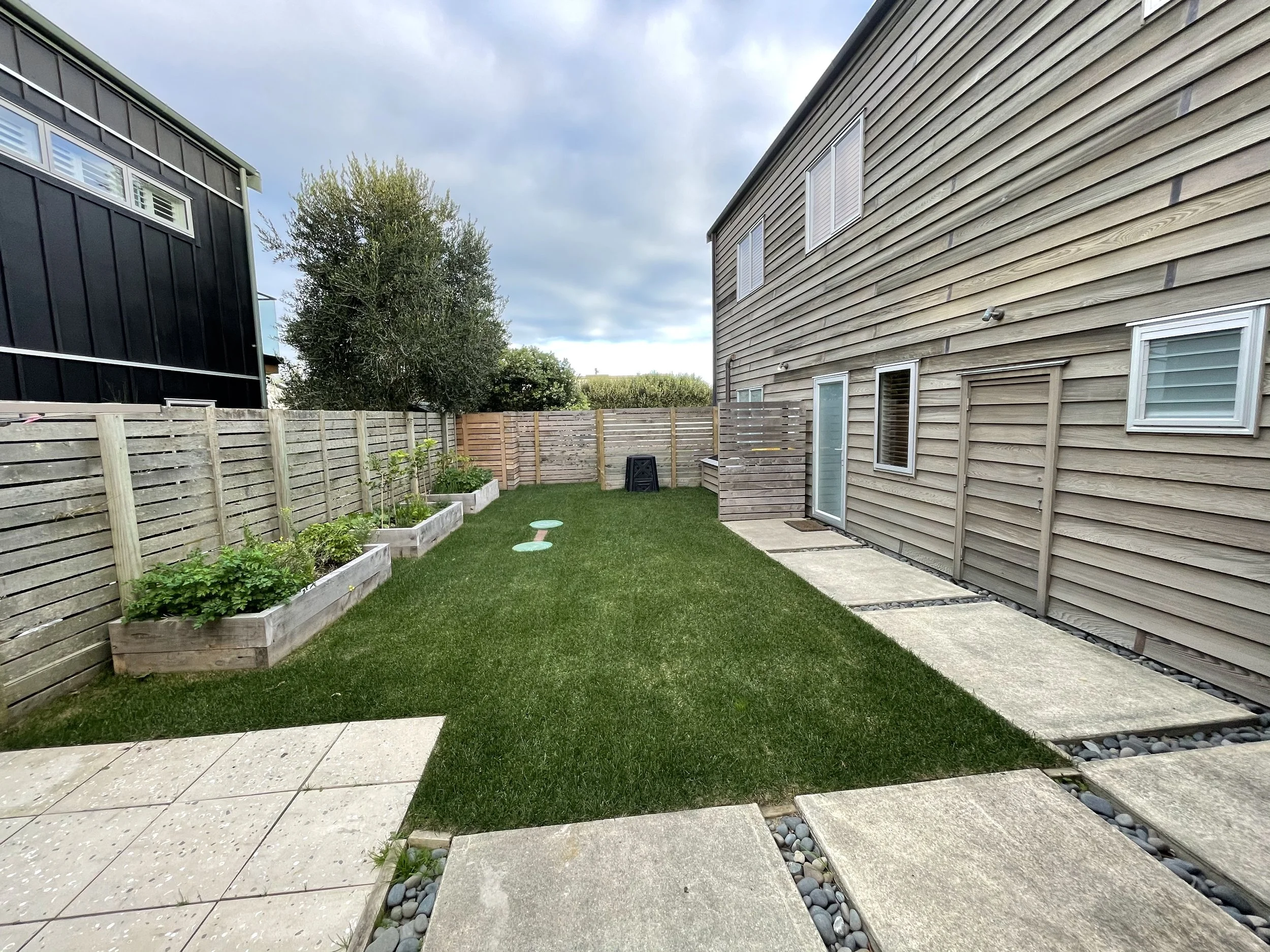 View of a backyard with a green lawn, raised garden beds, a wooden fence, and a beige house siding. A large tree is in the background, and there is a black building on the left. The sky is cloudy.