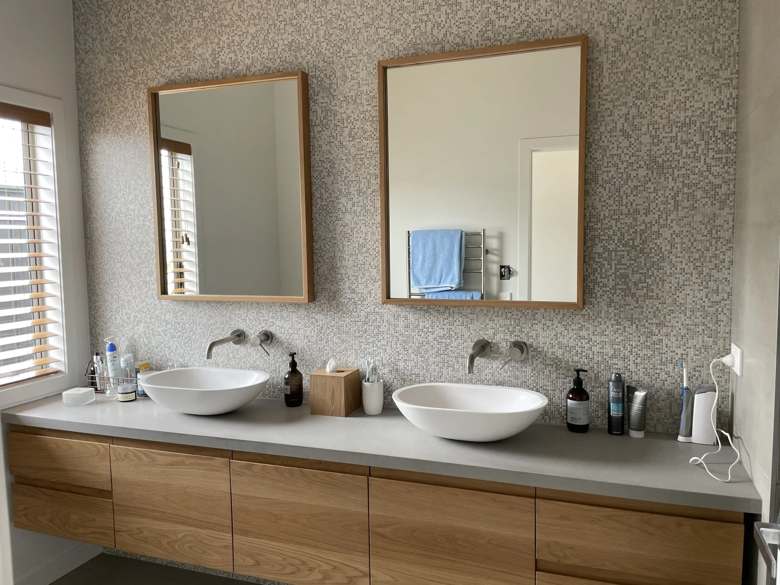 Bathroom with two white vessel sinks on a white countertop, wooden cabinet below, two rectangular mirrors with wooden frames, mosaic tile backsplash, window with blinds, and various toiletries on the countertop.