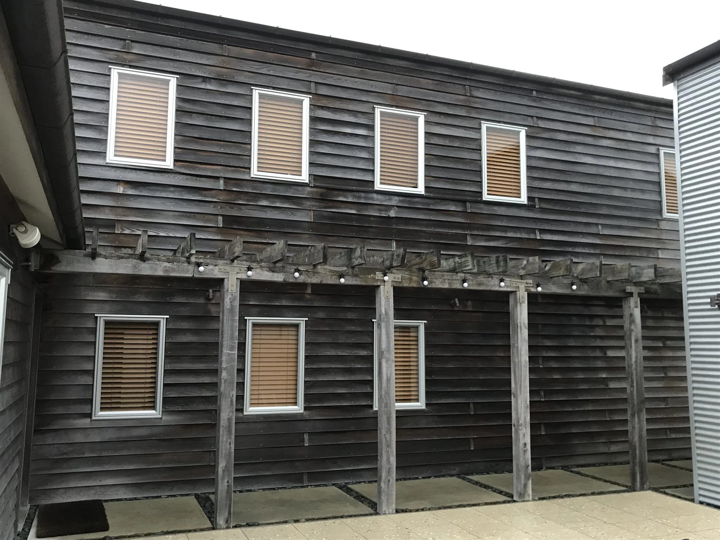 The photo shows the exterior of a modern building with dark wooden siding and multiple rectangular windows with brown blinds. The building has a sloped roof, and part of a porch with a wooden support structure and string lights hanging underneath is 